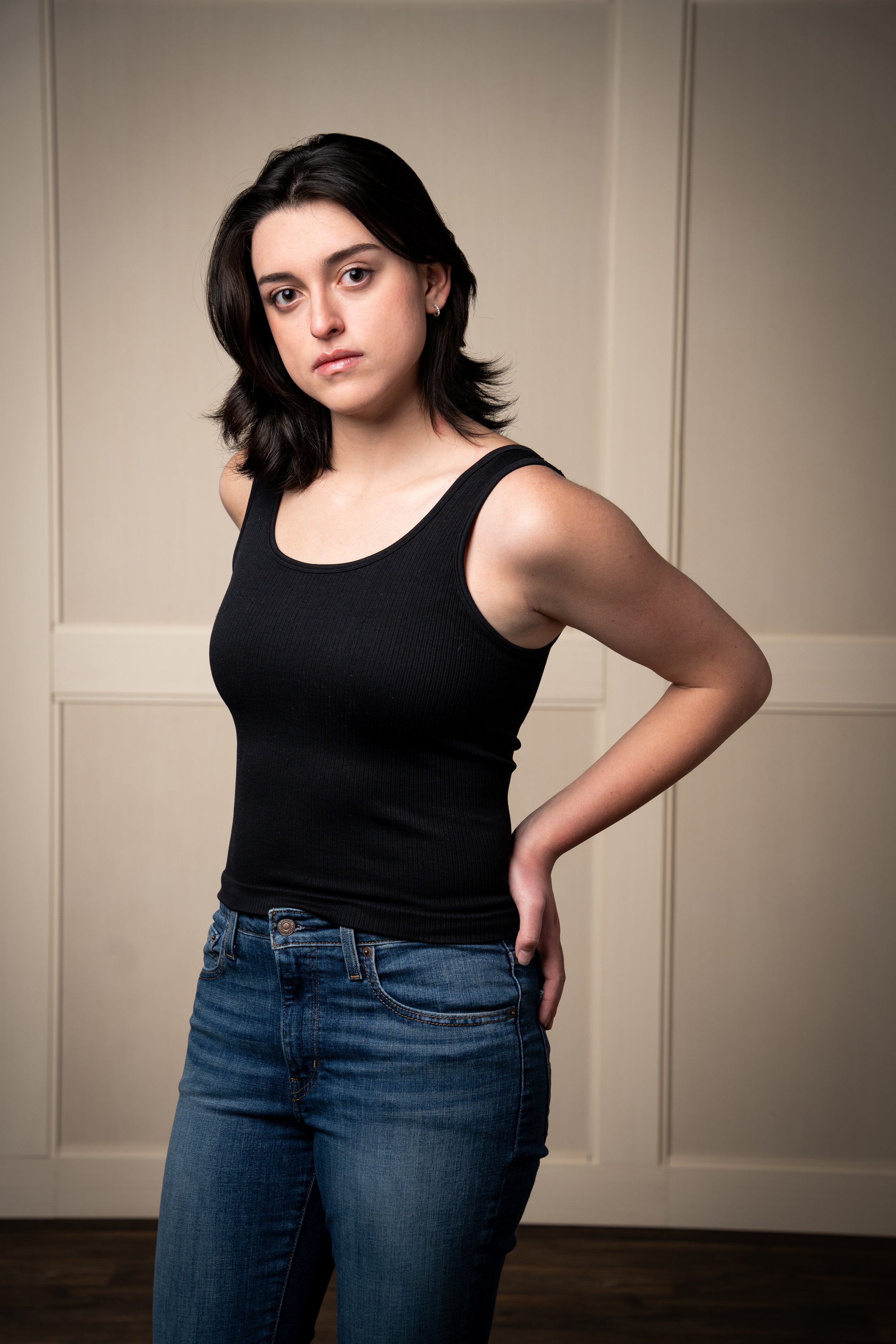 A young woman with dark hair wearing a black tank top and blue jeans standing in front of beige cabinets with a serious expression.
