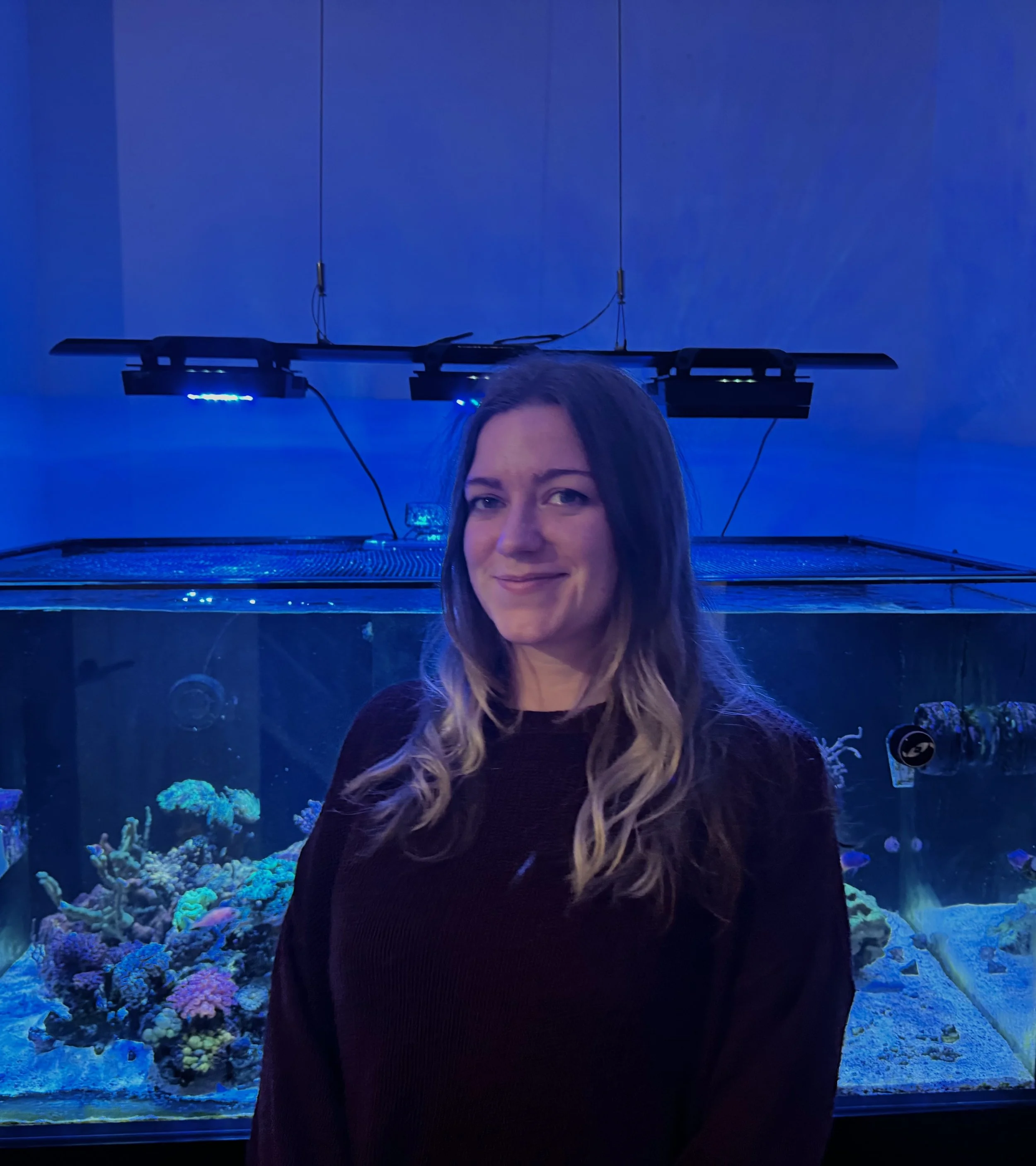 A woman with long, wavy hair and a dark sweater standing in front of an illuminated aquarium filled with colorful coral and various fish.