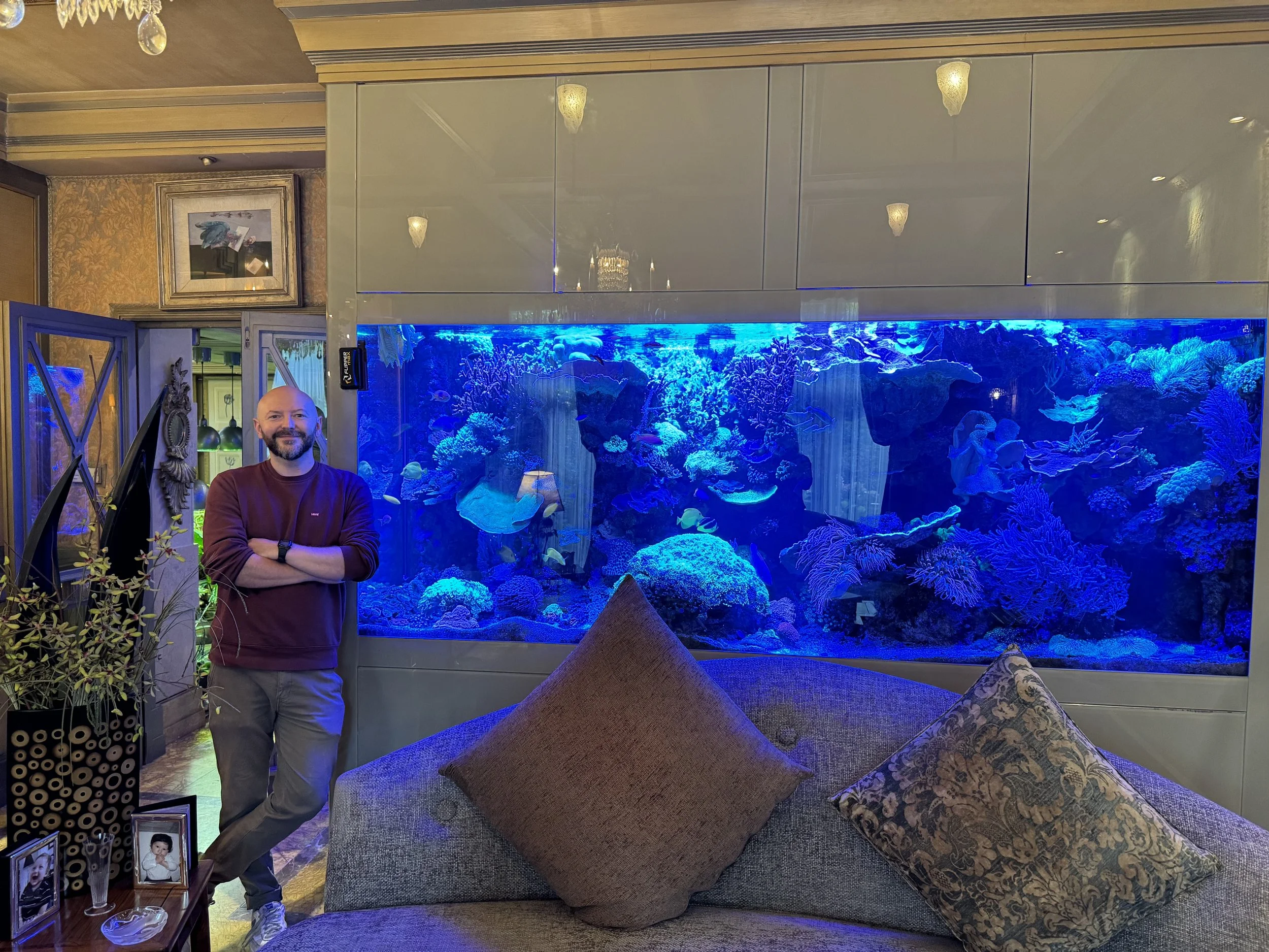 A man with a beard stands in front of a large aquarium filled with colorful coral and fish in a hotel lobby or lounge area. The background shows warm lighting and decorative wall art.
