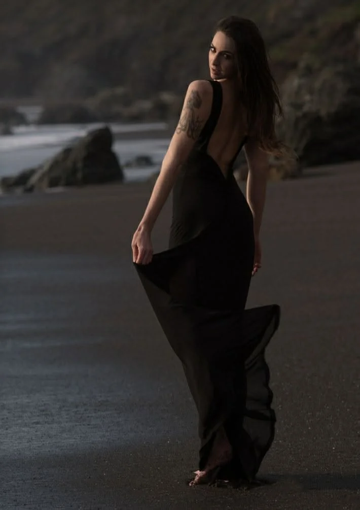A woman in a black dress standing on a beach with rocks and the ocean in the background at sunset.