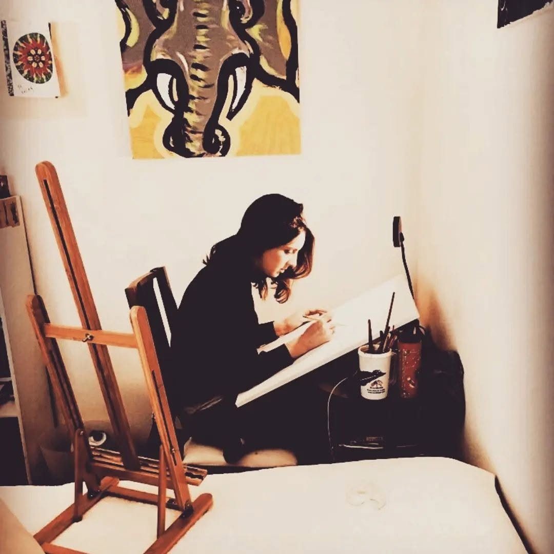 A woman sitting at a desk, working on a drawing or painting, surrounded by art supplies, with a colorful zebra painting on the wall behind her.