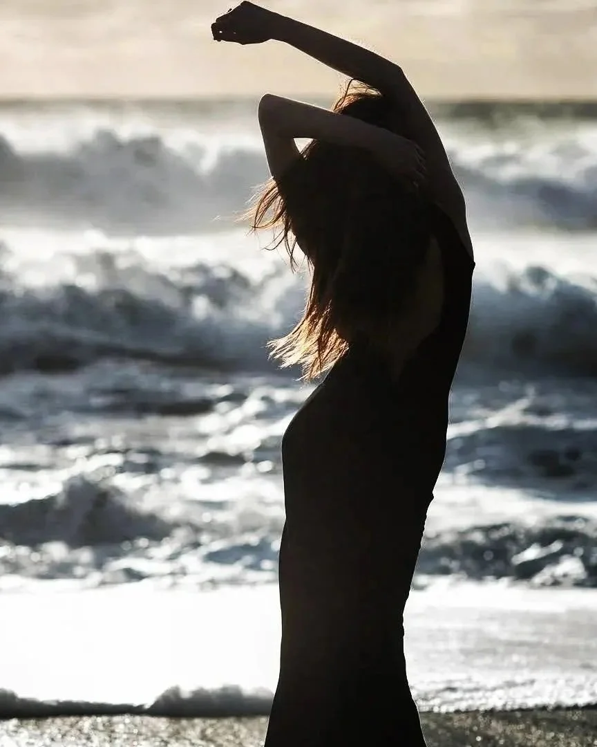 Silhouette of a woman with long hair standing on a beach, stretching her arms above her head, near the ocean
