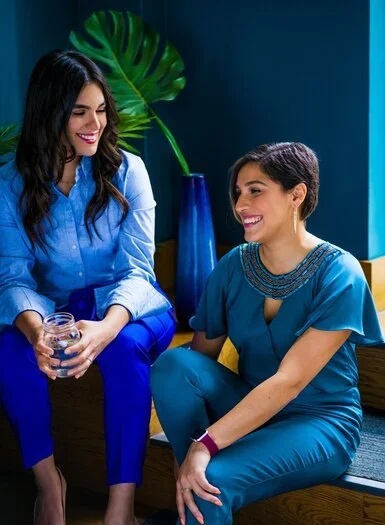 Two latina women smiling and talking indoors, one holding a glass of water, with a large leafy plant and blue decor in the background.