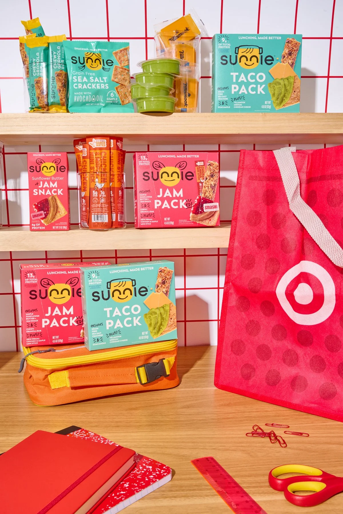 Shelf with Suncie snack packs, a Target reusable shopping bag, notebooks, scissors, a ruler, paper clips, a pencil case, and a red grid background.