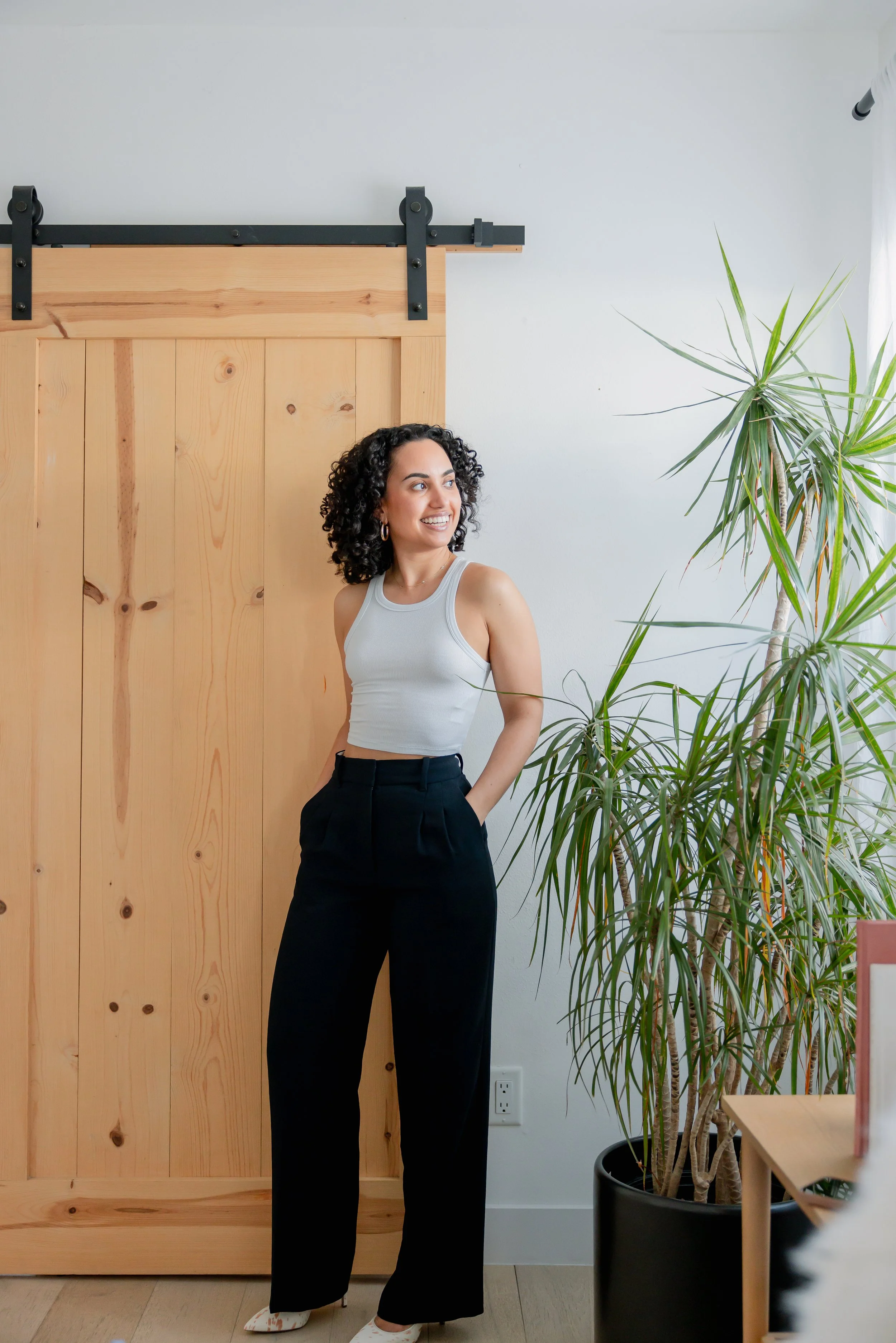 A young woman with dark curly hair, wearing a light gray sleeveless top and black high-waisted trousers, standing indoors in front of a wooden sliding barn door and a large green potted plant, smiling and looking slightly to the side.