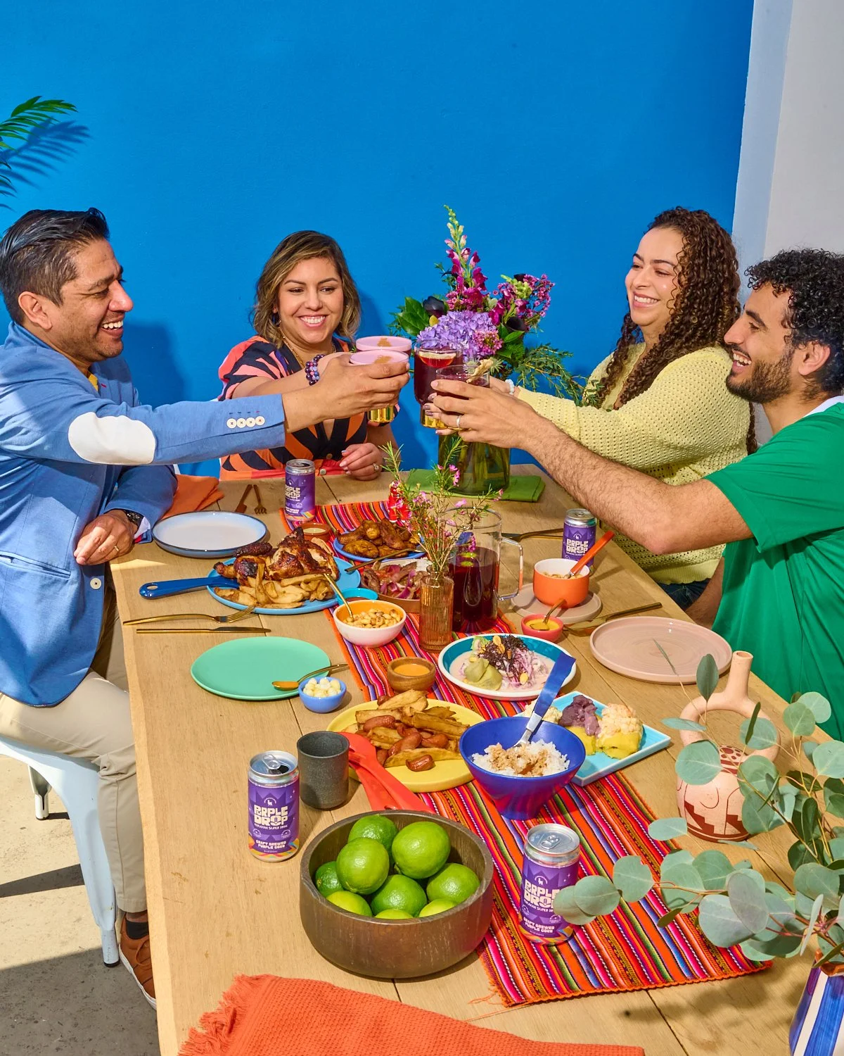 A group of five people smiling and raising glasses in a toast at a colorful dinner party, with a table full of food and drinks, and a blue background.