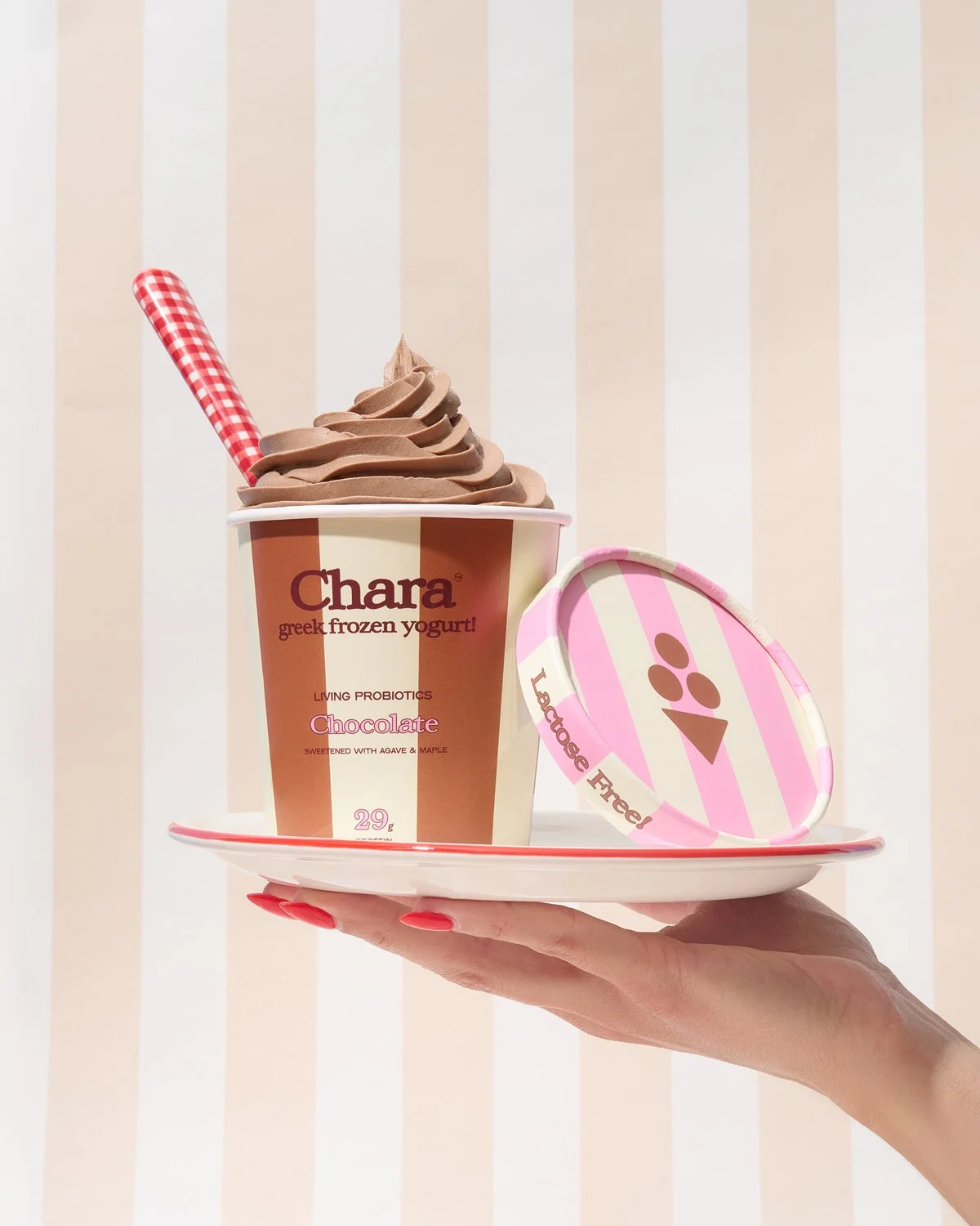 A hand holding a white tray with a cup of chocolate frozen yogurt, a pink-and-white striped lid, and a pink striped straw against a background of vertical beige and white stripes.