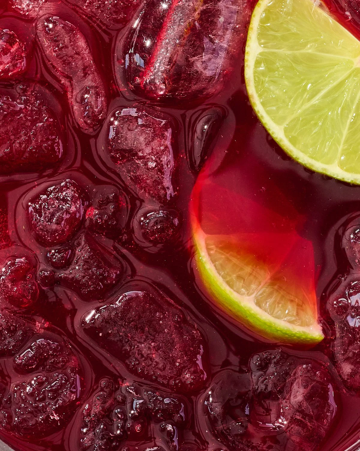 Close-up of a glass of iced beverage with ice cubes, a slice of lemon on the rim, and a vibrant red liquid.