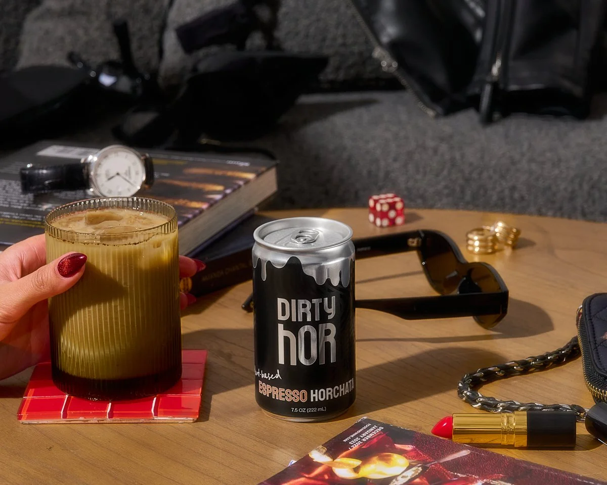 A hand with red nails holding a glass of iced coffee on a red tile coaster, on a wooden table with various items including a can of Dirty Hor Espresso Horchata, a wristwatch, sunglasses, a red domino, gold rings, a black chain, a lipstick, a backpack, an open book, a black bag, and a small purse in the background.