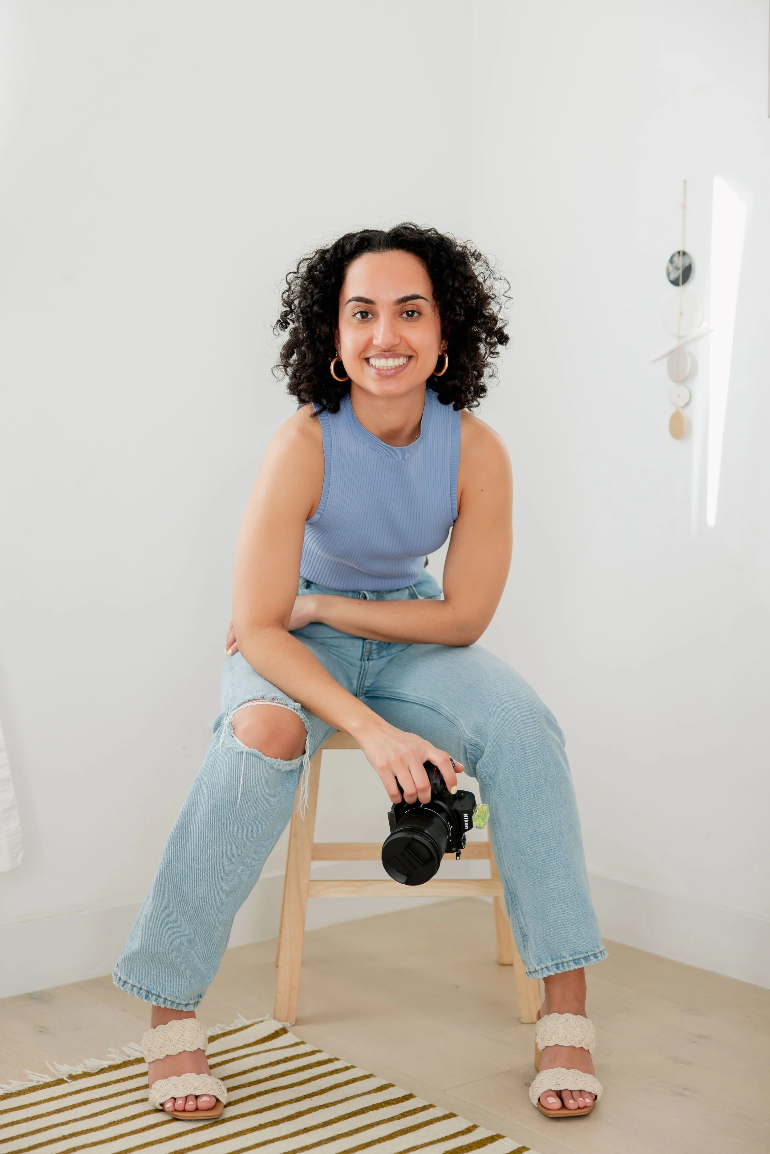 A smiling woman with curly black hair, wearing a blue sleeveless top and ripped jeans, sits on a wooden stool with a camera in her hand. She is in a minimalistic room with light-colored walls, and a striped rug on the floor.