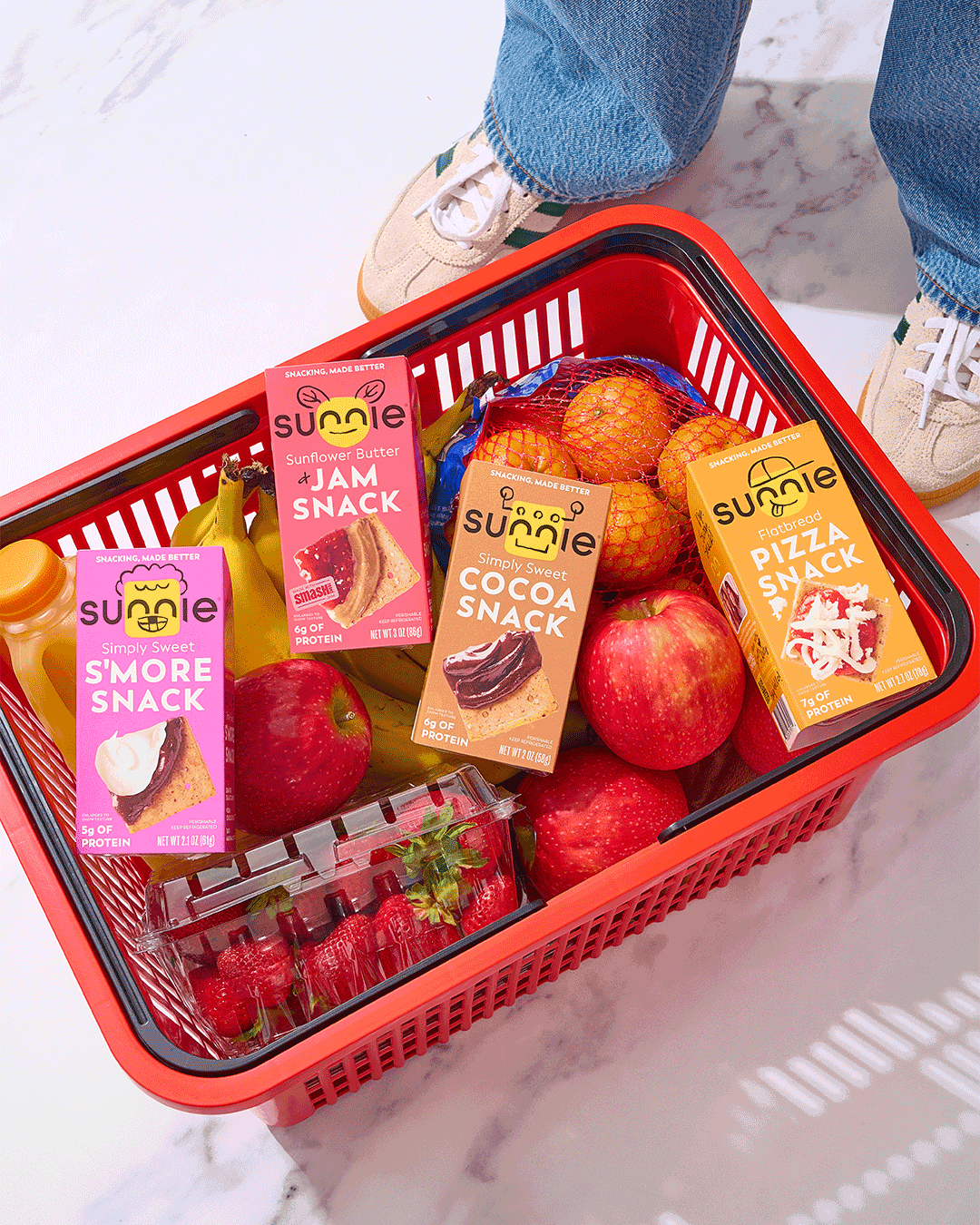 Red shopping basket filled with apples, strawberries, a bag of lemons, and three snack boxes labeled Sunlie: S'more Snack, Sunflower Butter & Jam Snack, Cocoa Snack, and Flatbread Pizza Snack.