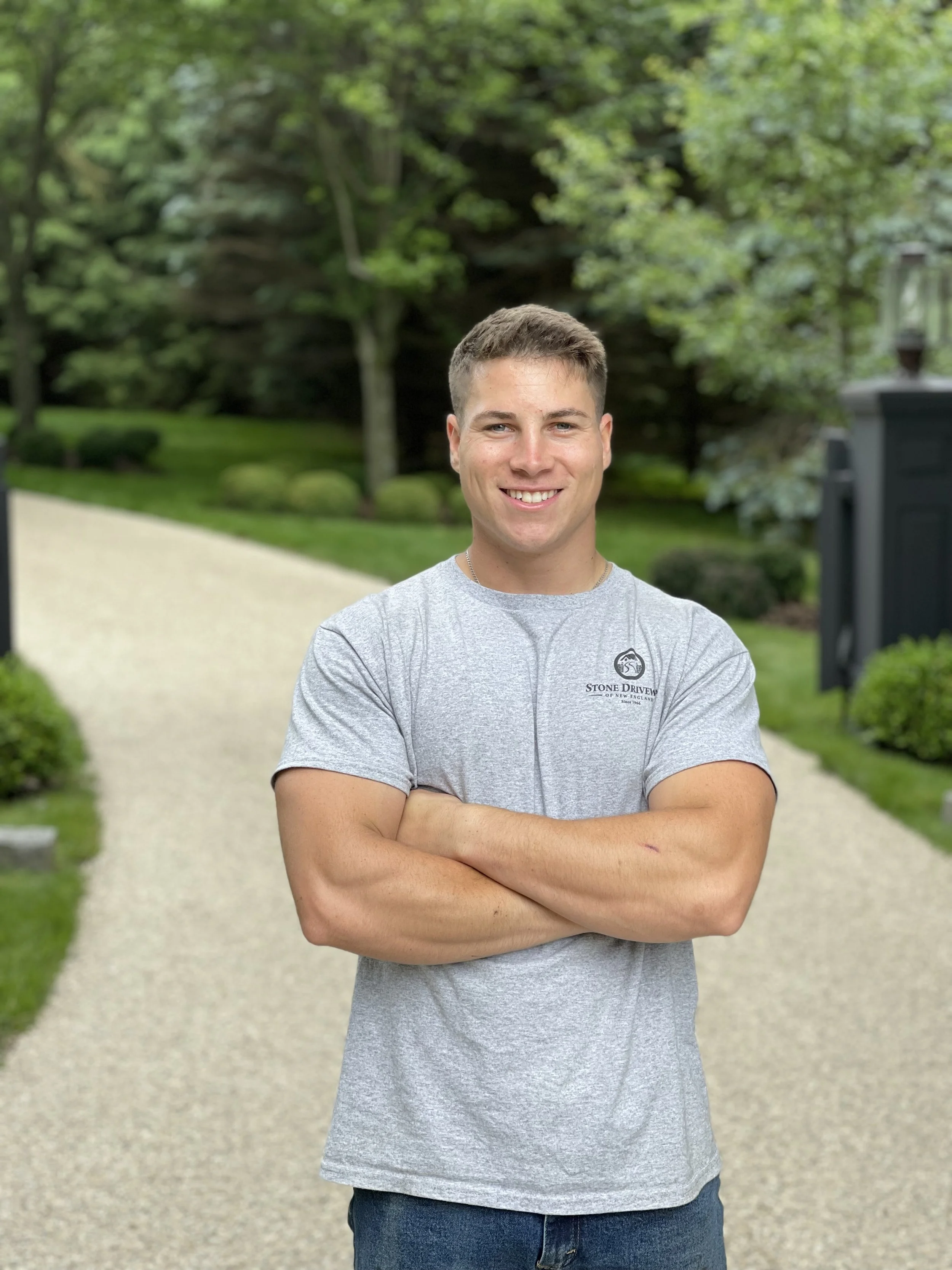 A young man with short brown hair smiling and standing outdoors with arms crossed. He is wearing a gray T-shirt and blue jeans. The background shows a gravel path, green trees, and bushes.
