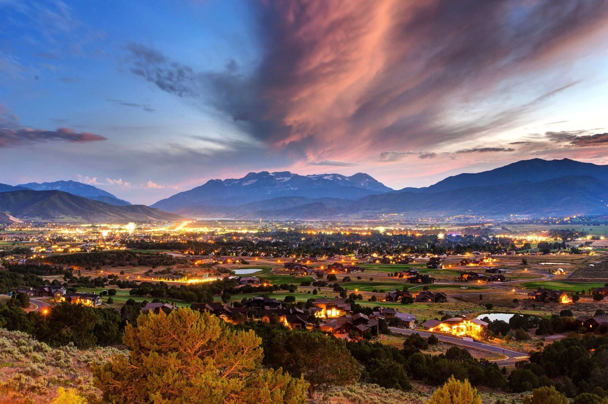 Scenic view of a valley with houses and city lights, with a mountain range in the background under a colorful sky at sunset or sunrise.