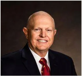 A professional headshot of a smiling man in a suit and tie against a dark background.