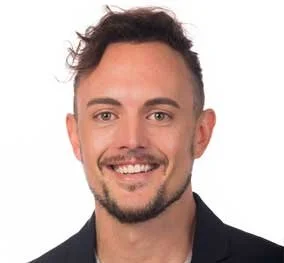 Headshot of a smiling man with short curly brown hair, light beard, wearing a black blazer and a light-colored shirt against a white background.