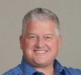 Headshot of a smiling middle-aged man with short gray hair, wearing a blue shirt, against a neutral background.