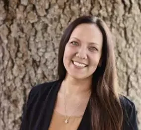 A woman with long brown hair smiling in front of a tree.