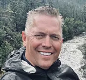 A man with short, gray hair smiling outdoors in front of a river and trees.