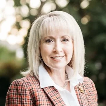A woman with blonde hair smiling outdoors, wearing a plaid blazer and white shirt.