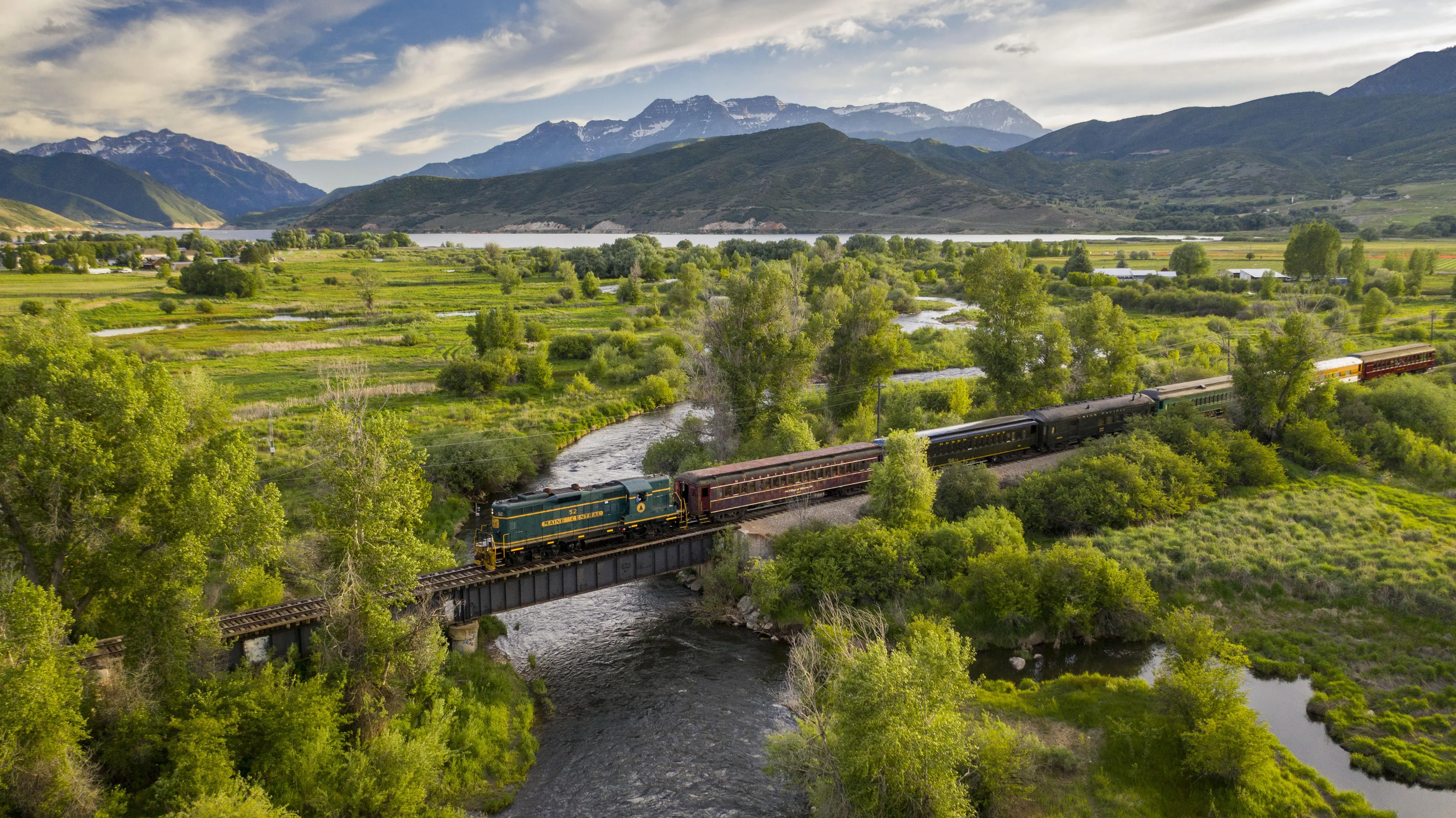 A scenic landscape with a mountain range in the background, a river, green fields, and a vintage train with black and green cars traveling over a bridge.