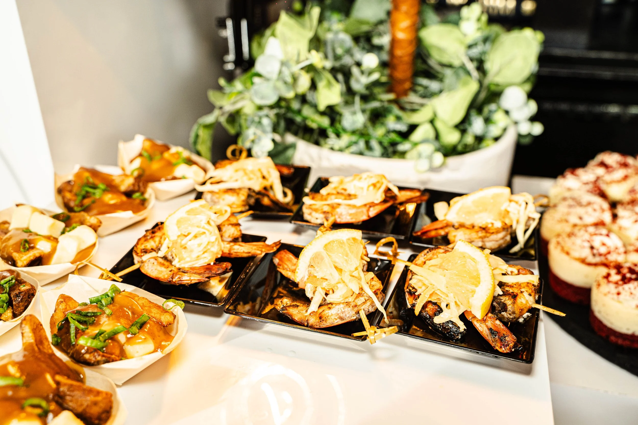 Close-up of a variety of appetizers on black square plates, including grilled shrimp topped with lemon slices, and small bowls of meat topped with sauce and chopped green onions, with a large green leafy plant in the background.