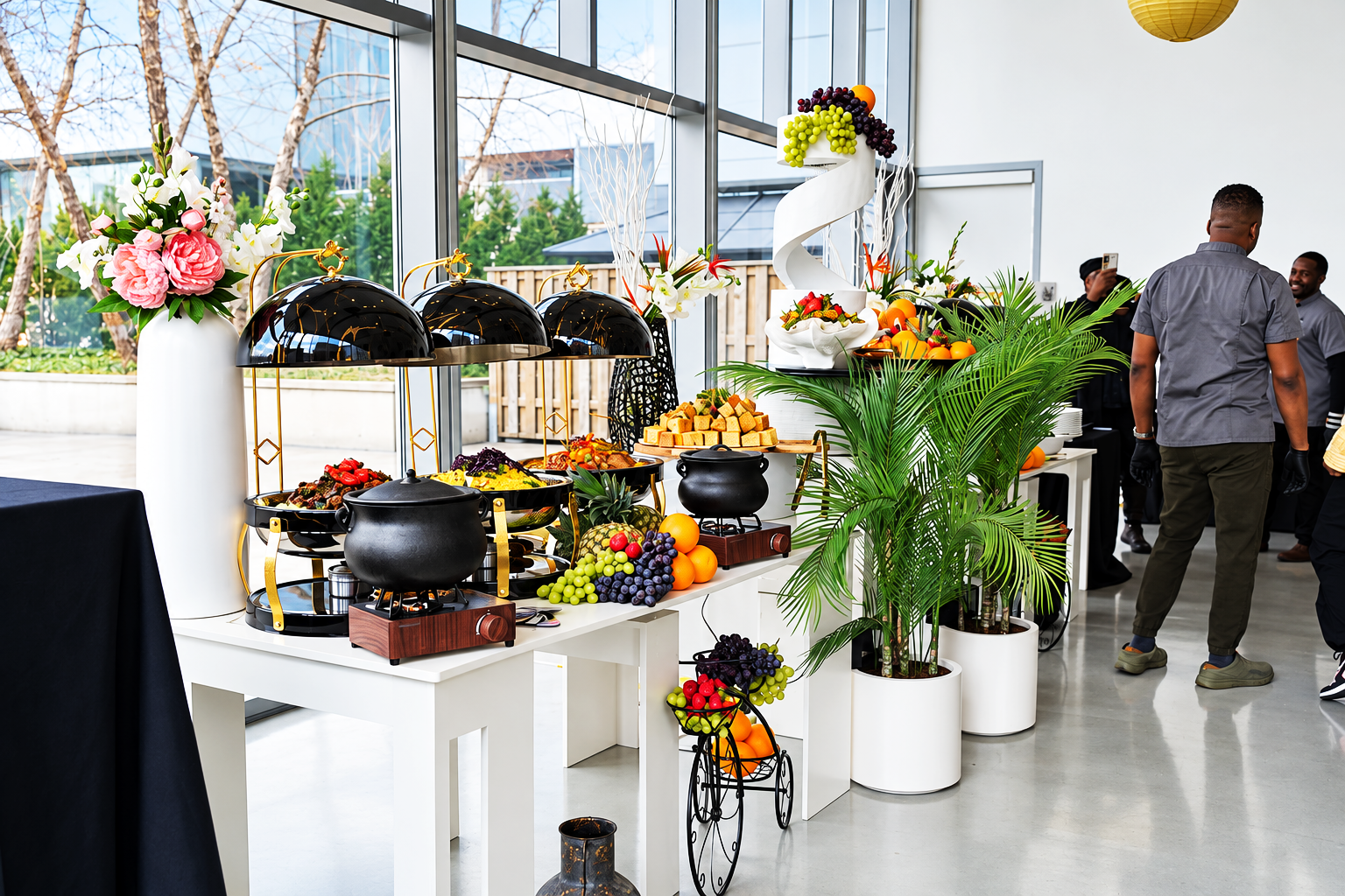 A buffet-style setup with food and decorative elements, including a large white flower vase, black chafing dishes, fresh fruit, and greenery, inside a bright, modern space with large windows.