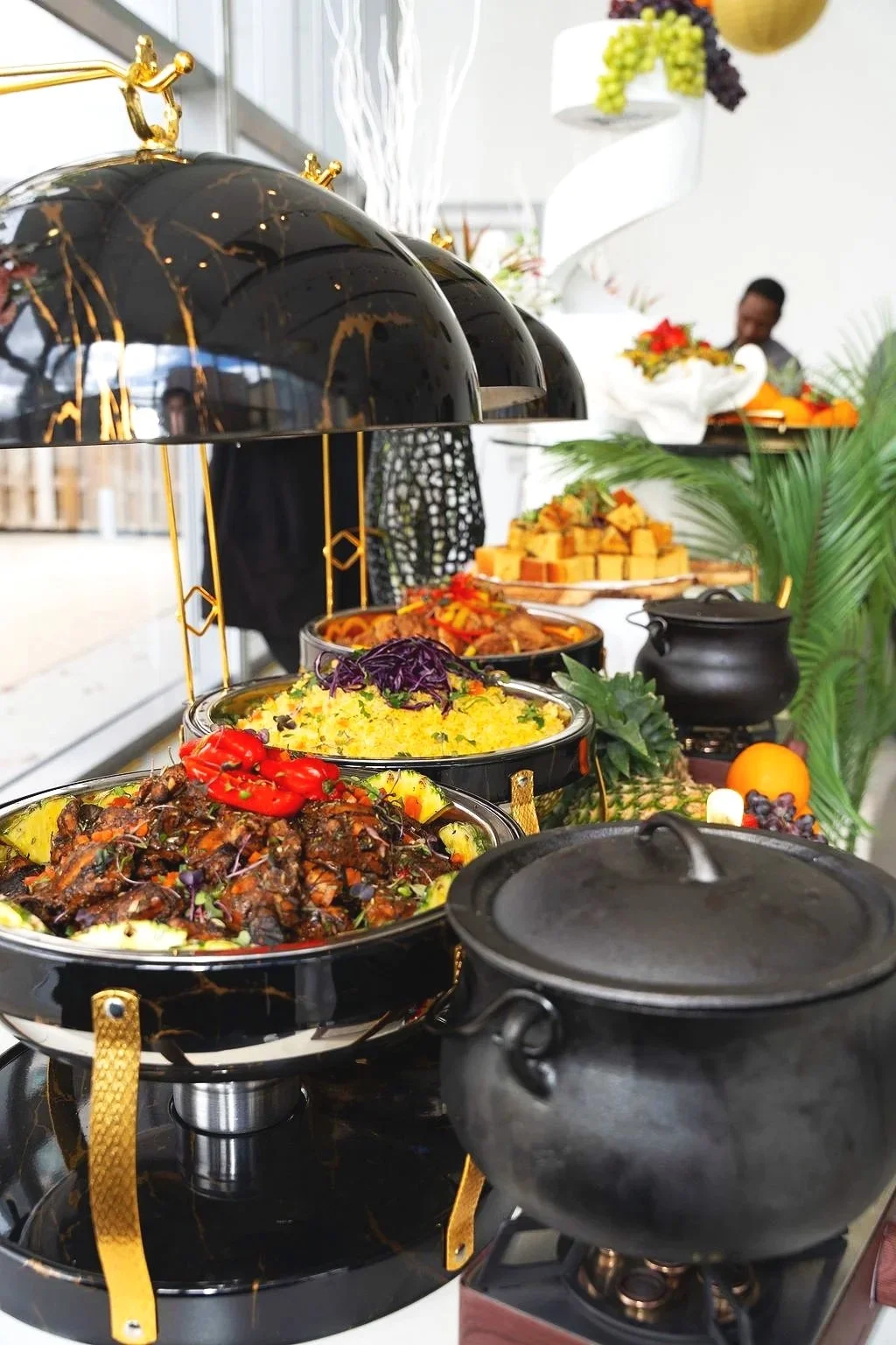 Buffet spread with various hot and cold dishes, including vegetables, meats, and desserts, set on a table with decorative greenery and a person in the background.