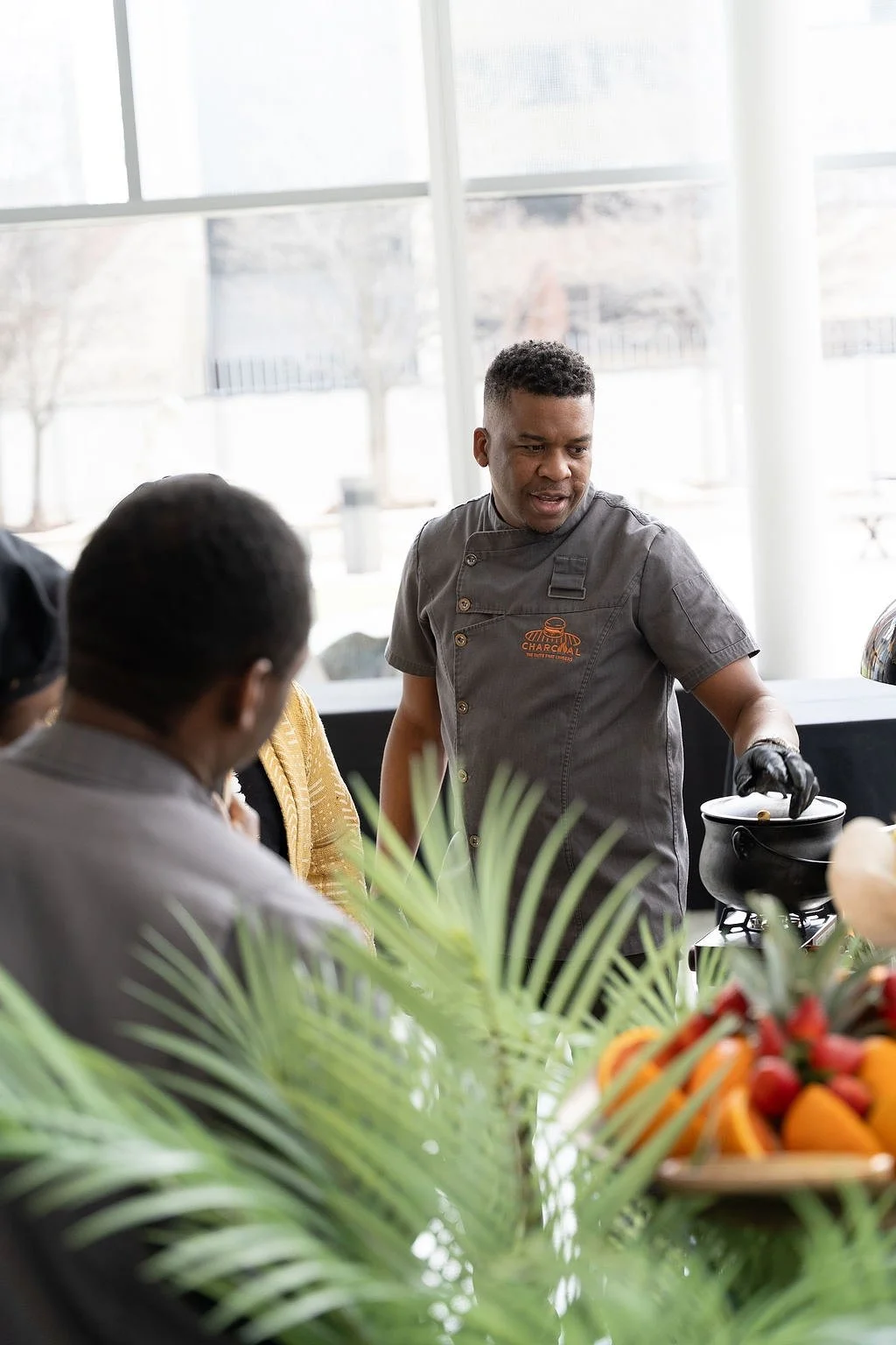 A chef in a gray uniform with logo, cooking at a table with attendees in a bright, modern room with large windows, plants, and a basket of fruits
