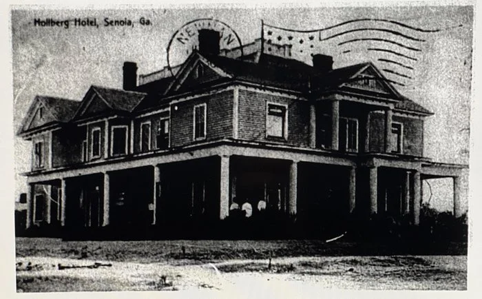Black and white historical photograph of the Holberg Hotel in Senoia, Georgia, showing a large two-story wooden building with a wrap-around porch, several windows, and a gabled roof, with a postal cancellation mark partially visible on the top right.