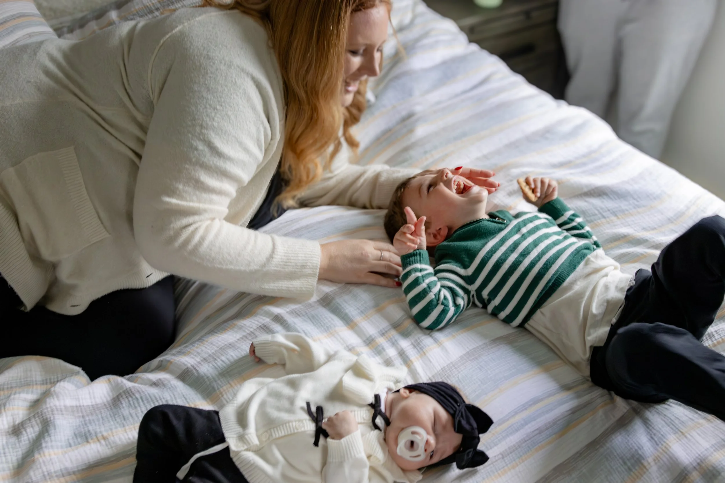 A woman with red hair lying on a bed, smiling and looking at a young boy with dark hair, who is also lying on the bed and laughing. The boy has a pacifier in his mouth and is wearing a green and white striped sweater.