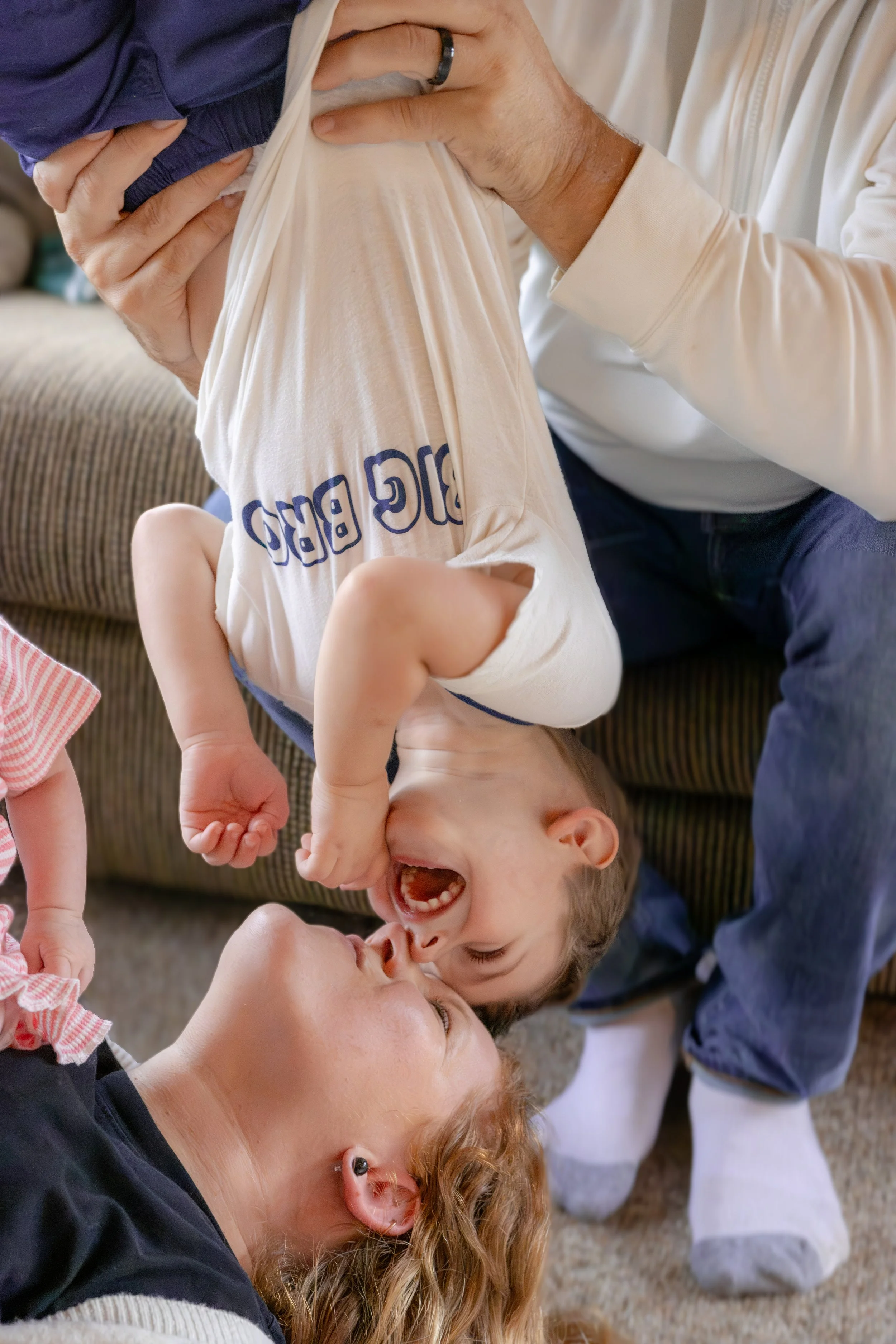 A young boy hanging upside down, laughing while playing with his mother and sibling, lying on the floor. They are indoors, on a carpeted floor.