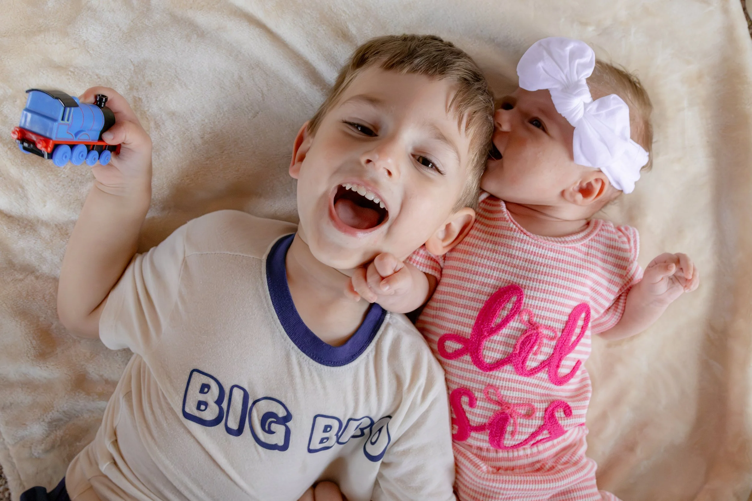 Two children lying on a soft beige blanket, the older boy smiling with his mouth open, holding a blue toy train, and the younger girl wearing a large white bow headband and a pink and white striped shirt, resting close to him.