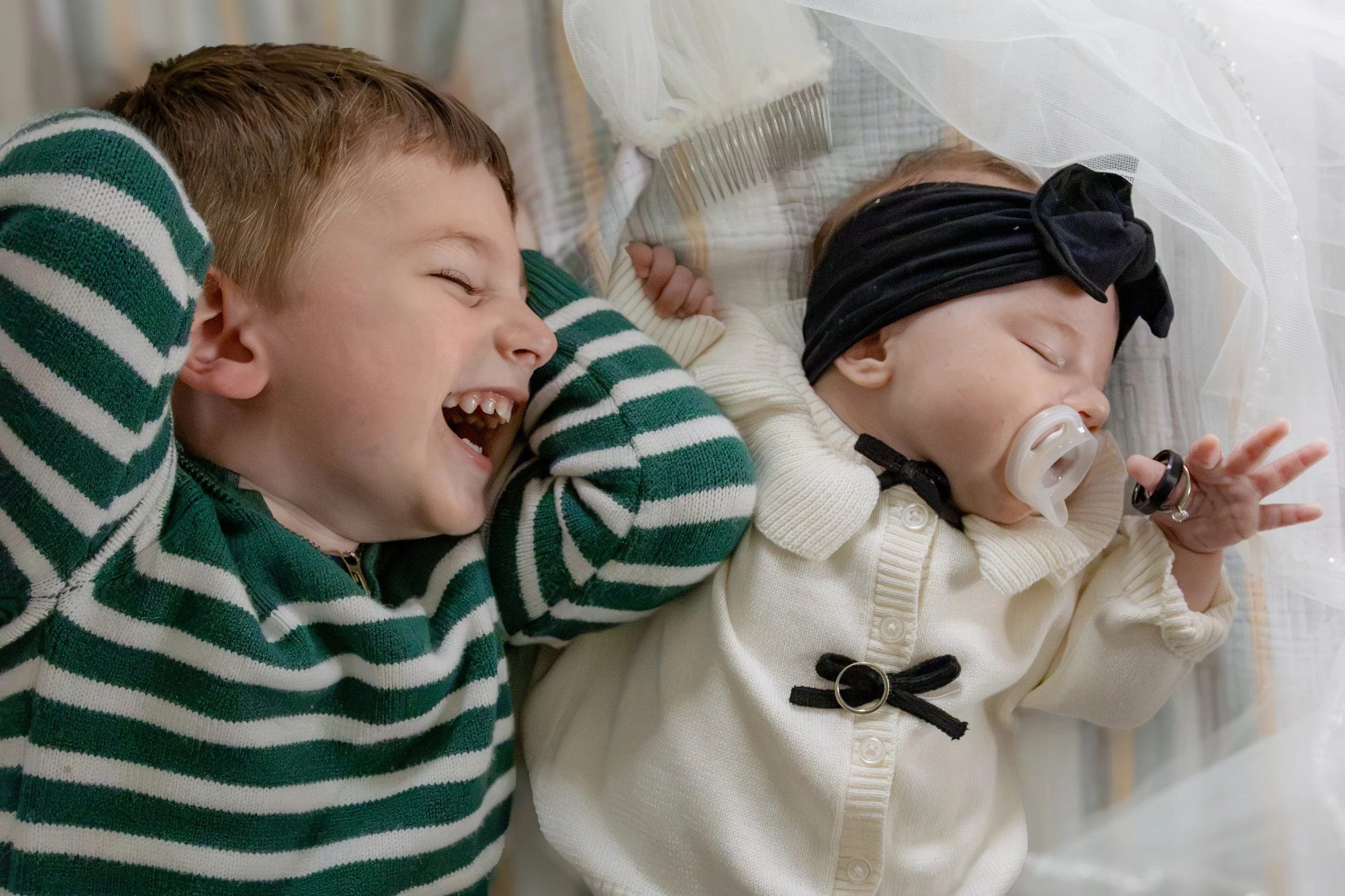 Two young children lying on a bed, smiling and sleeping. One child is wearing a striped green and white sweater, the other in a light-colored cardigan and a black headband, with a pacifier and an IV in the hand.