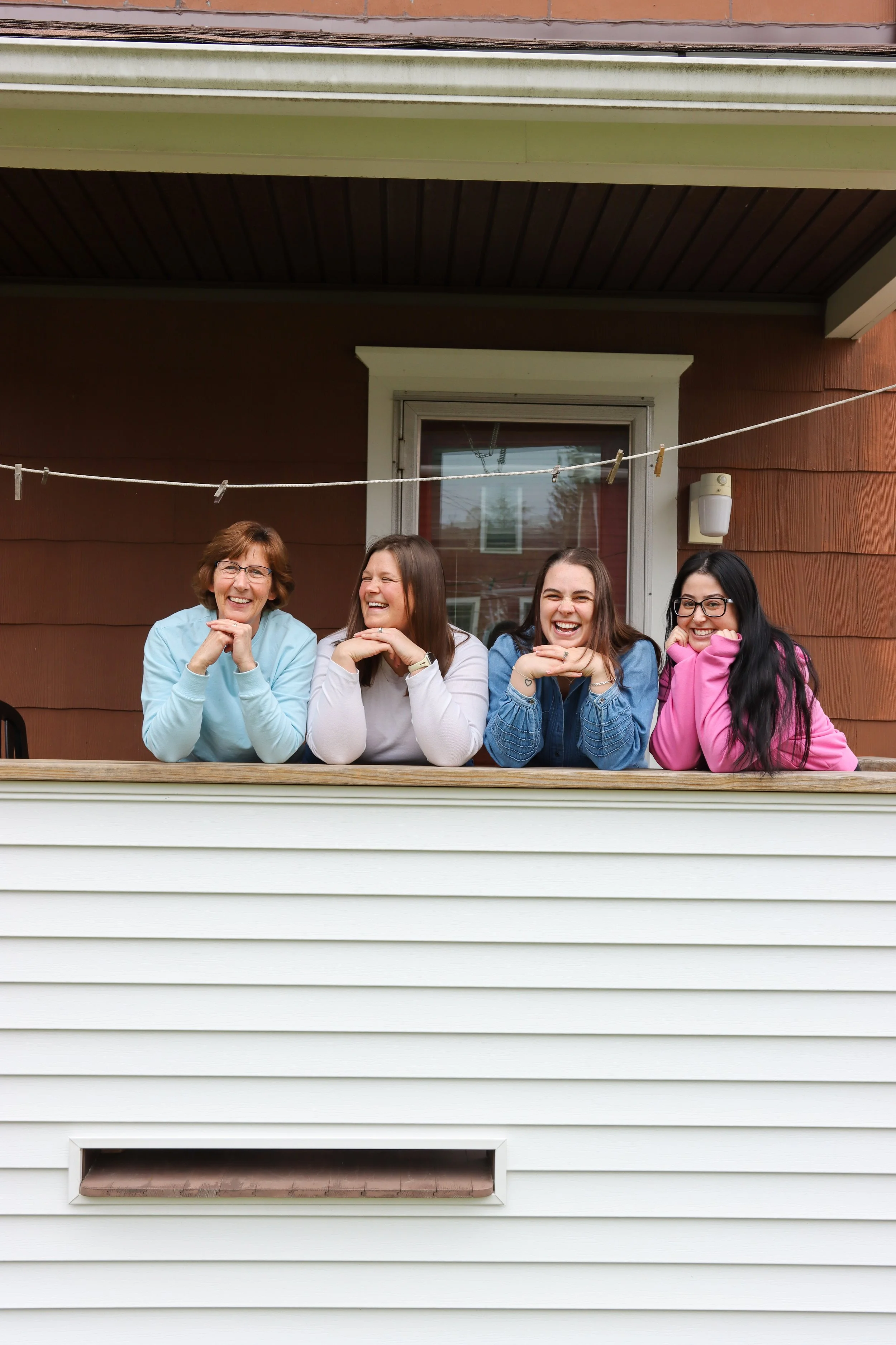 Four women sitting on a balcony, laughing and smiling, with a clothesline above them, a window behind, and brown and white exterior house siding.