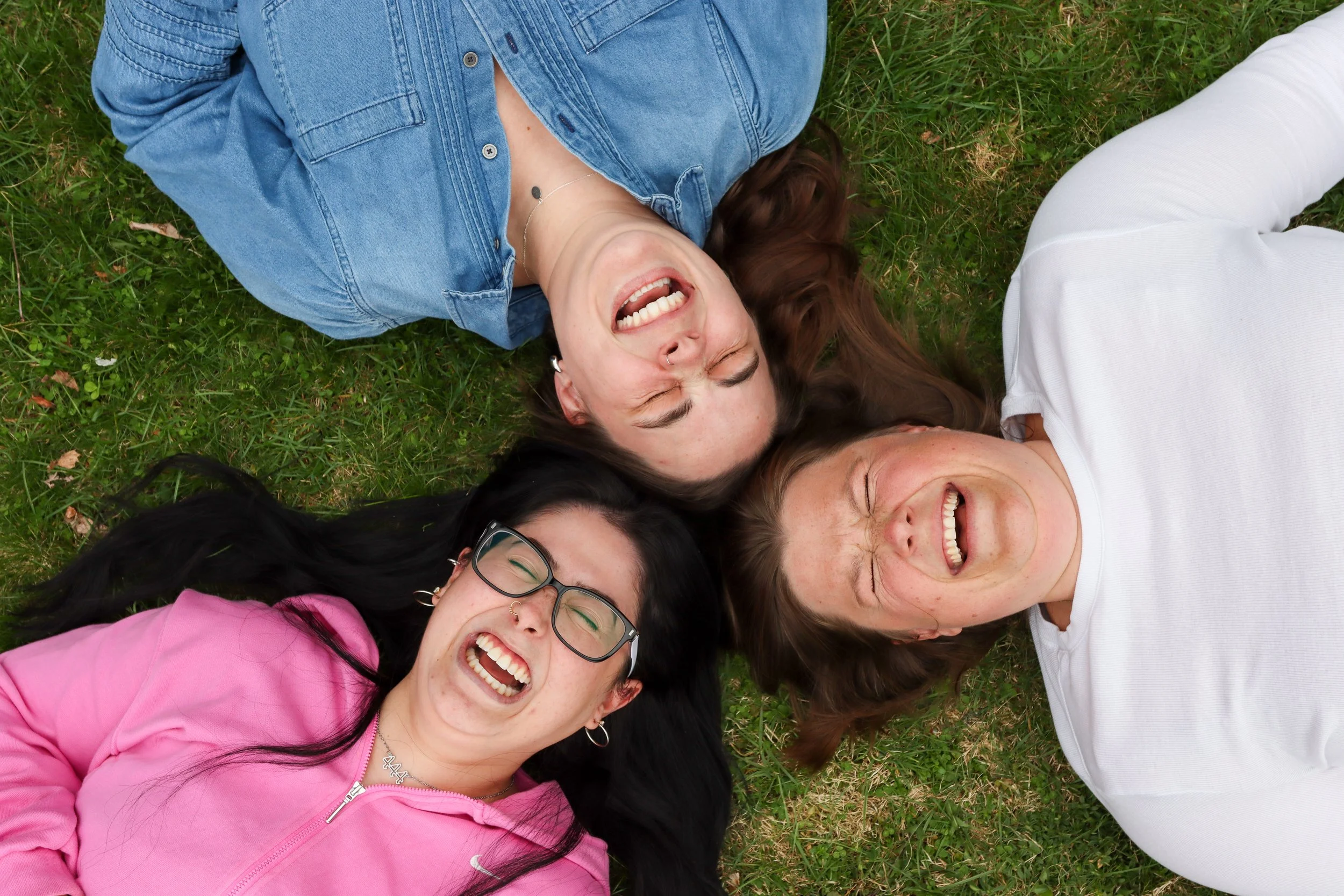 Group of smiling women lying on grass with their heads close together, showing happiness and friendship.