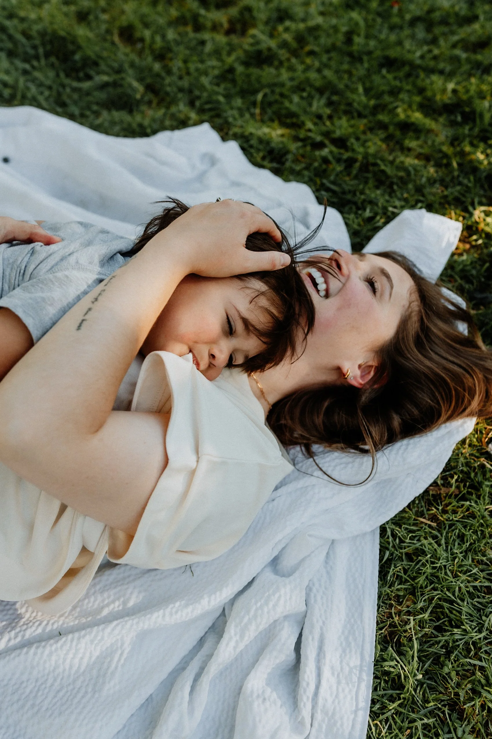Two women lying on a white blanket on the grass, hugging and smiling outdoors.