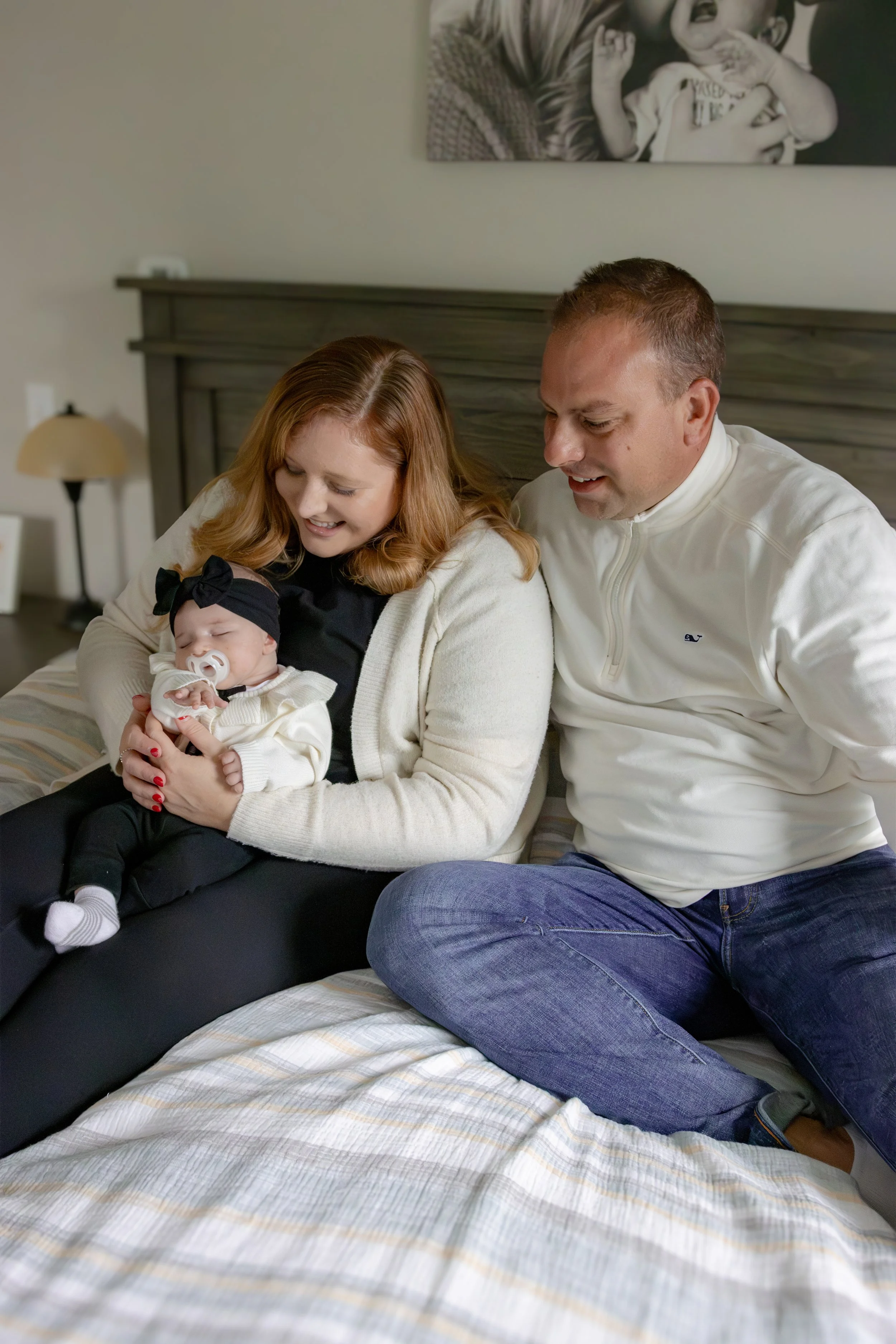 A family sitting on a bed: a woman holding a baby with a pacifier, a man sitting next to them, all smiling and looking at the baby