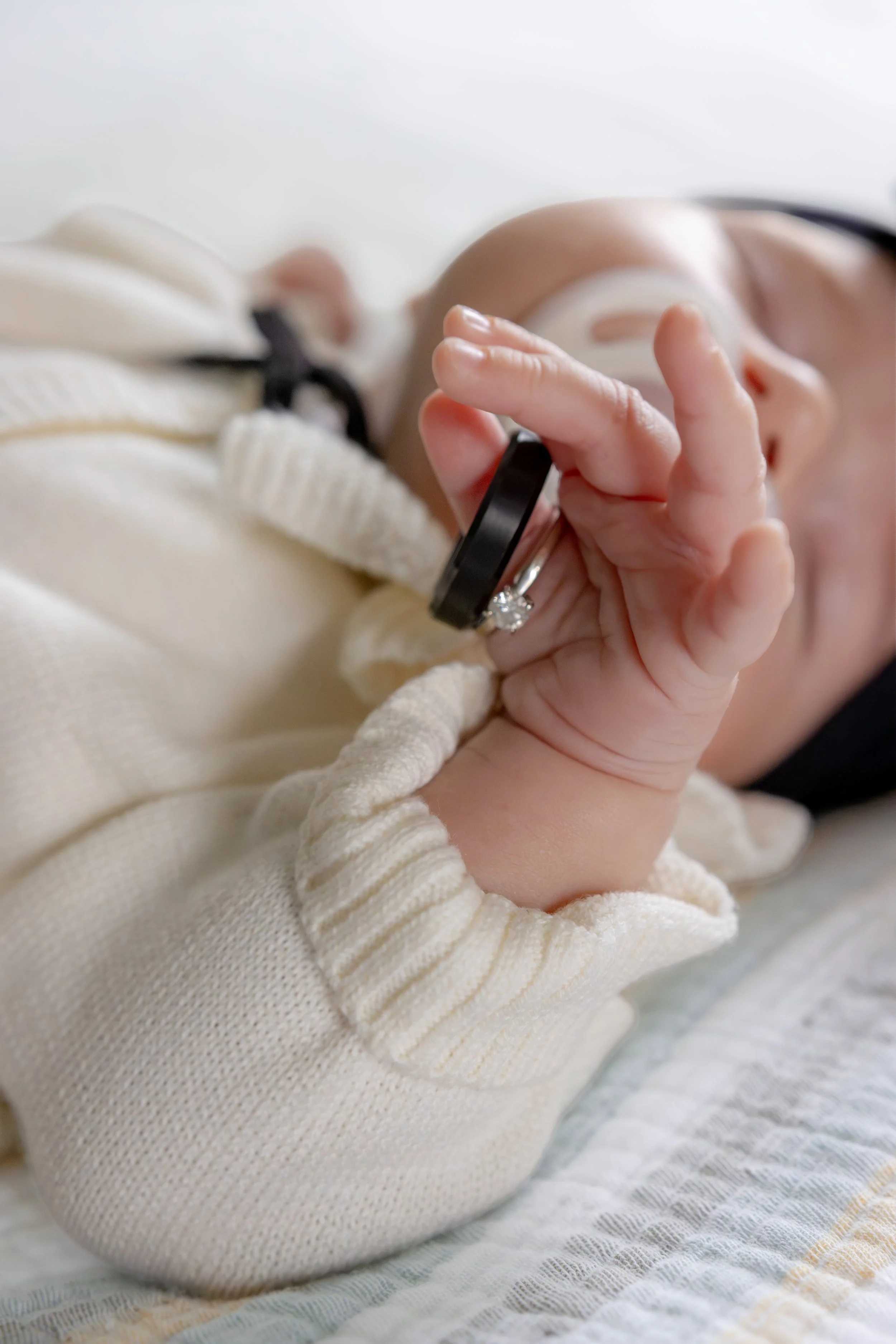A close-up photo of a baby holding mom and dad's wedding rings. The baby is wearing a cozy, cream-colored sweater, and their face is partly visible but out of focus.