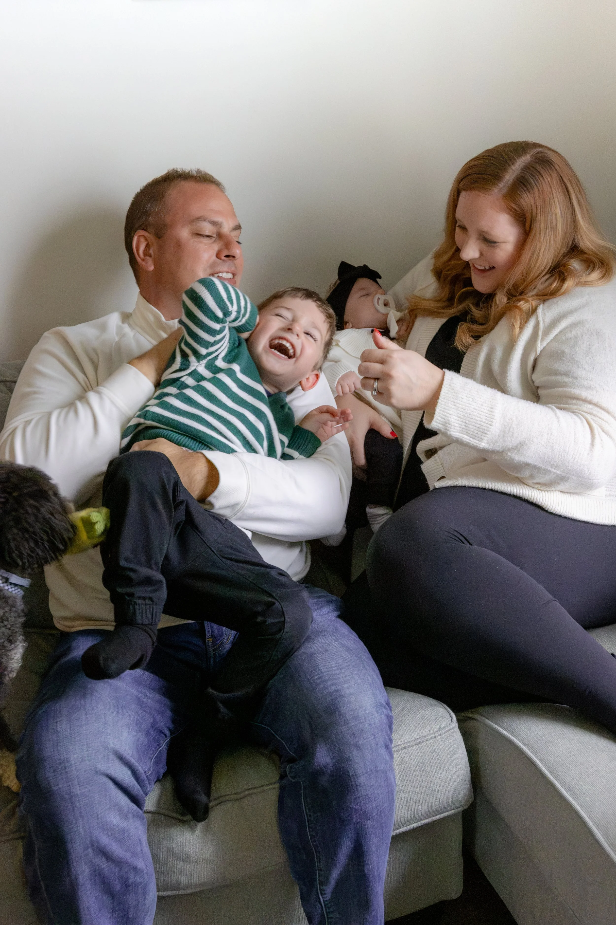 A family with two young children sitting on a couch, celebrating a special occasion indoors. The father is holding a laughing toddler, and the mother is holding a newborn baby. They are all smiling and enjoying the moment together.