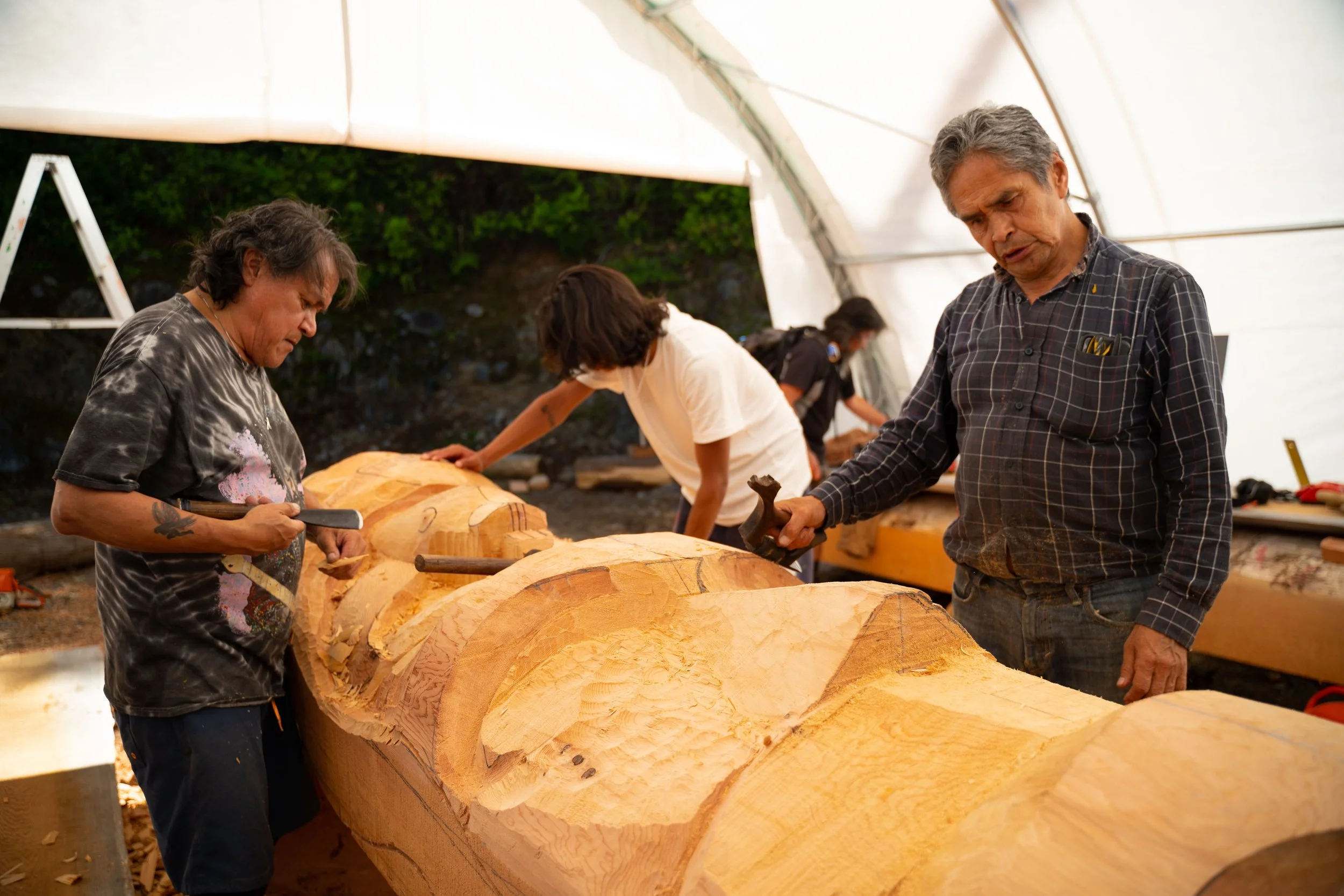Three individuals working on a totem pole inside a tent. They are using tools to carve and detail the wood.