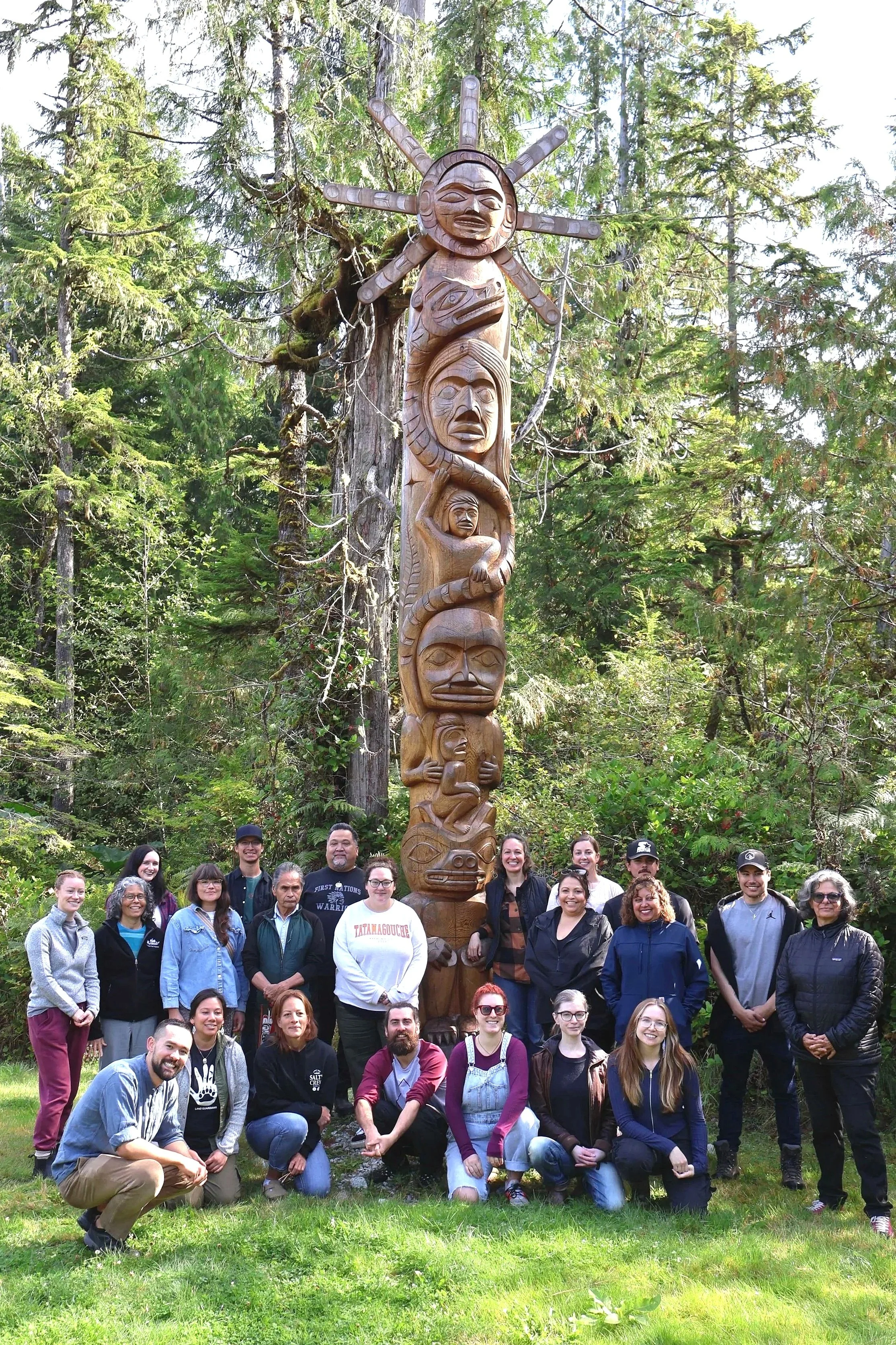 Group of diverse people standing in front of the NaWas totem pole at the Clayoquot Campus with forest trees in the background.