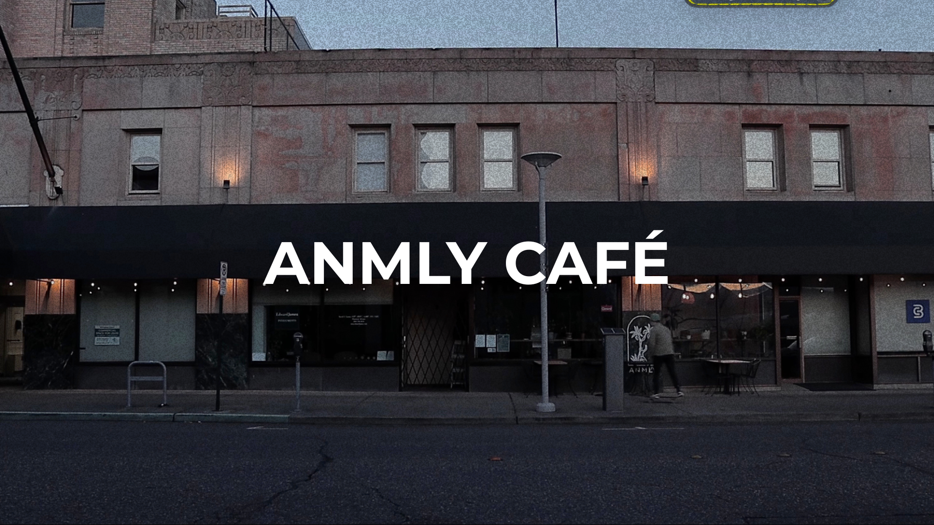 A building with a black awning that has white text reading 'ANMLY CAFÉ.' The exterior walls are light-colored with visible signs of wear and small windows. There are street lights, a lamp post, a parking meter, and a person walking on the sidewalk in front of the café, with some outdoor seating and string lights visible.