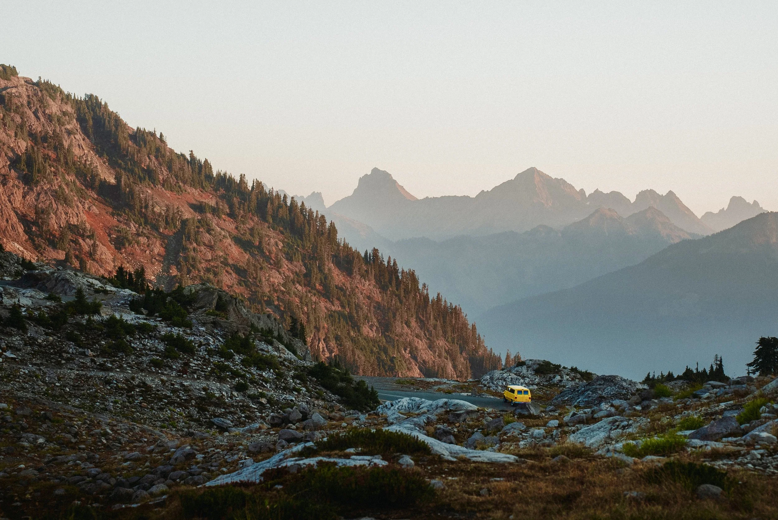 A yellow vehicle driving on a rocky mountain road with trees and mountains in the background during sunset.