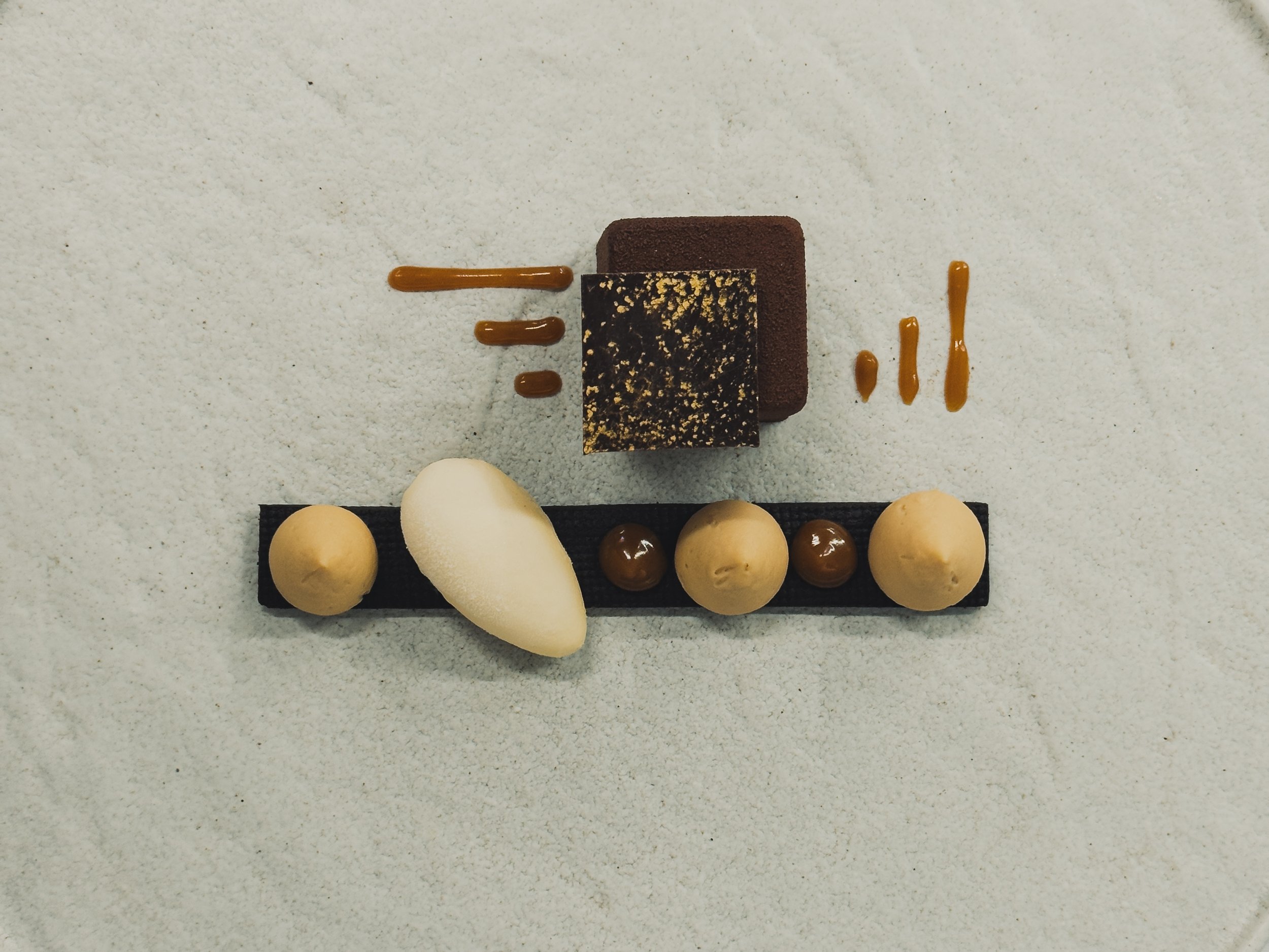 Assorted desserts including a chocolate mousse, white dome-shaped mousse, round scoops of ice cream or mousse, and caramel sauce on a white surface.