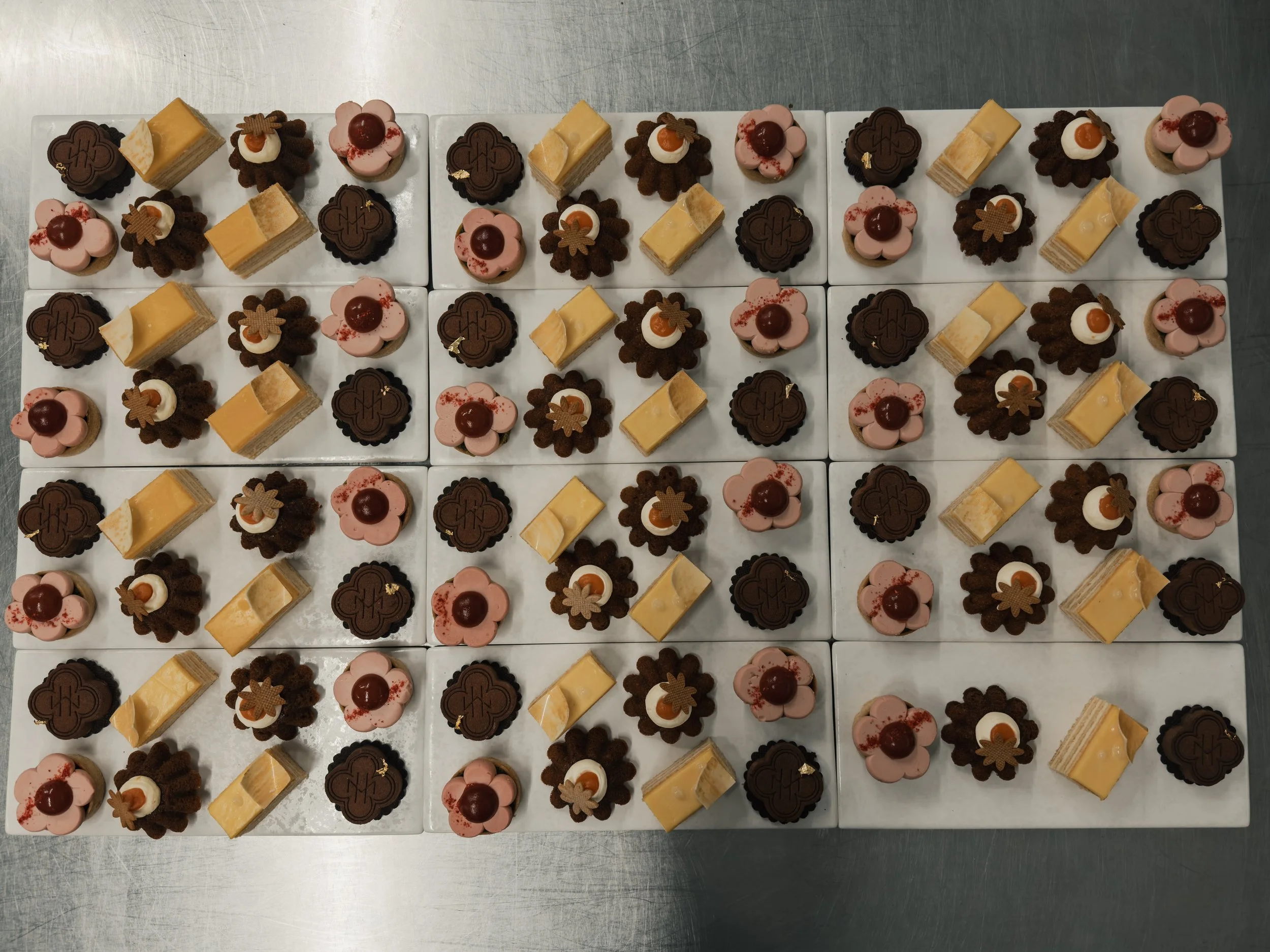 A tray of assorted petits fours includes pink flower-shaped cakes with a cherry on top, layered yellow cakes, and round chocolate cakes with a decorative design on top, arranged on white serving platters.
