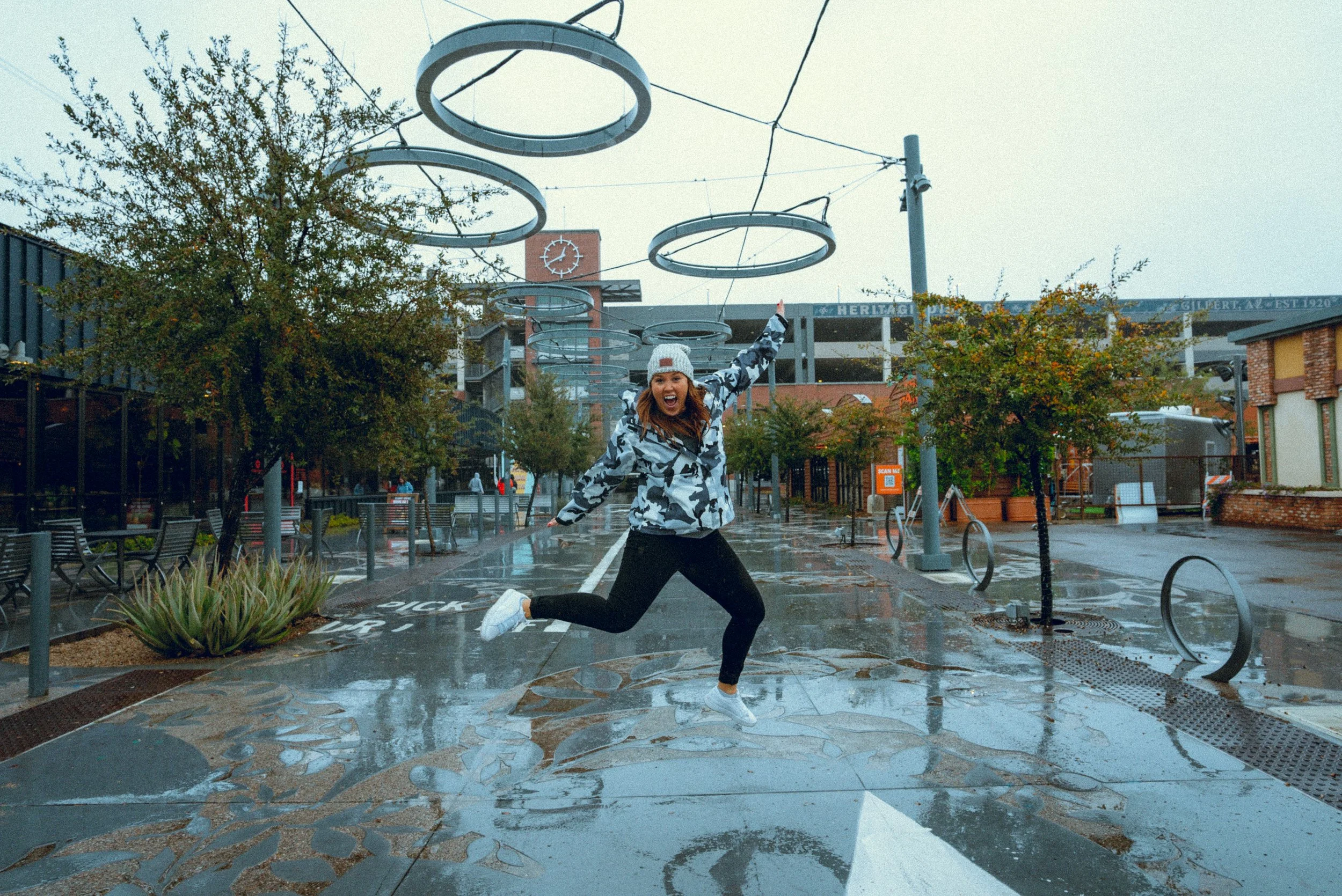A woman wearing a white beanie, camouflage jacket, and black pants is jumping with her arms raised and smiling in a wet outdoor urban area with some puddles, trees, benches, and buildings in the background.