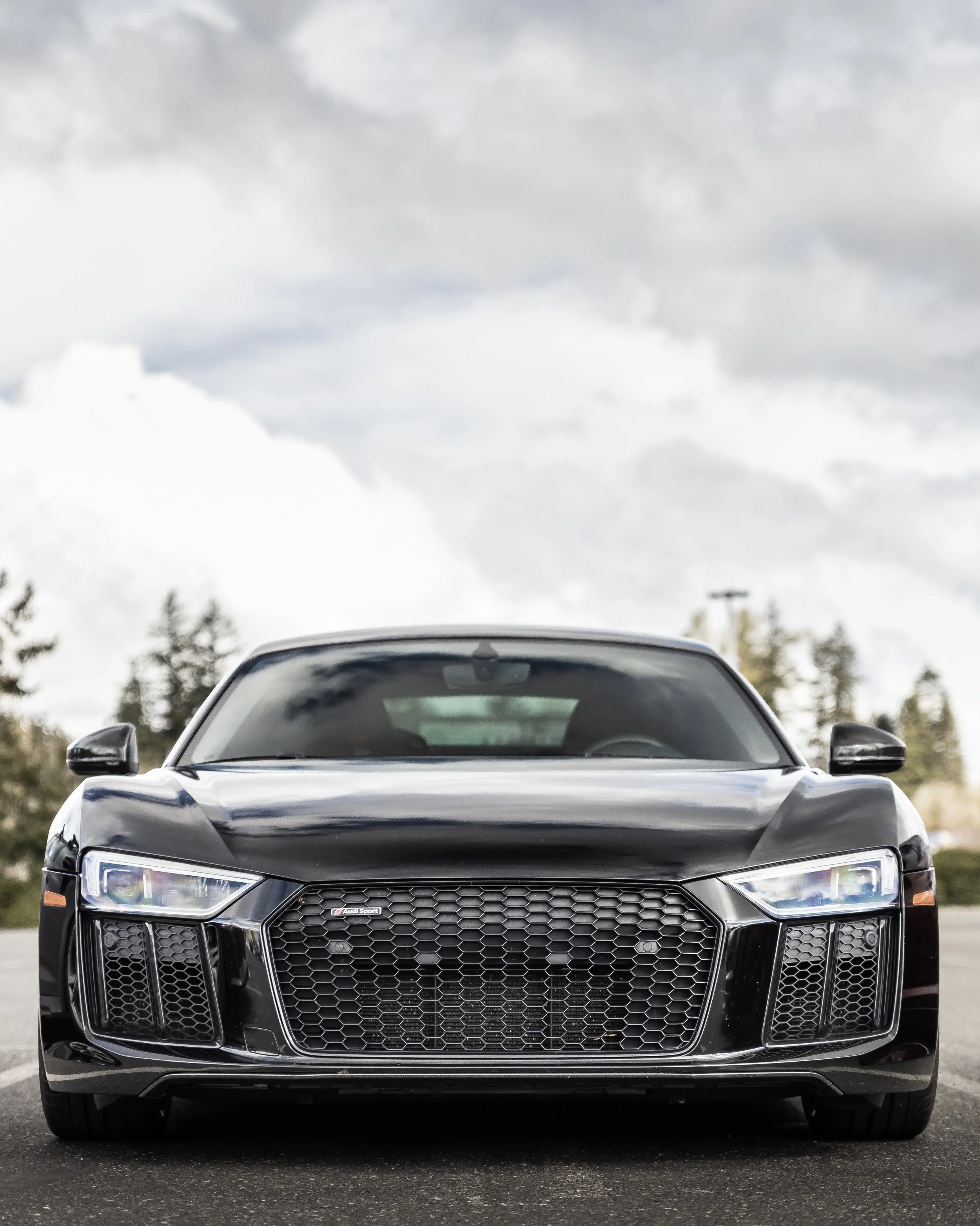 Front view of a black Audi Sport car parked on a wet surface with trees and cloudy sky in the background.