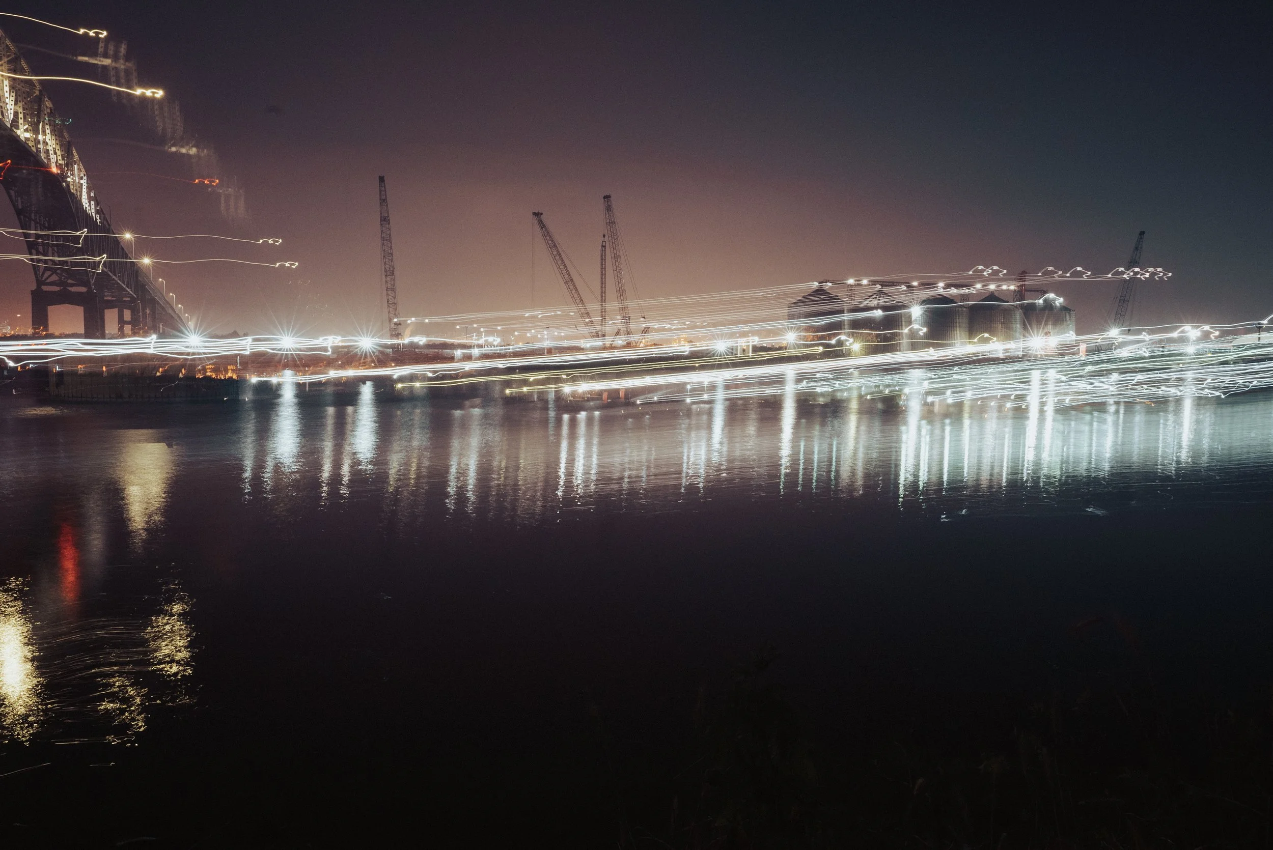 A night view of a bridge with light trails and reflections on the water, with construction cranes and buildings in the background.