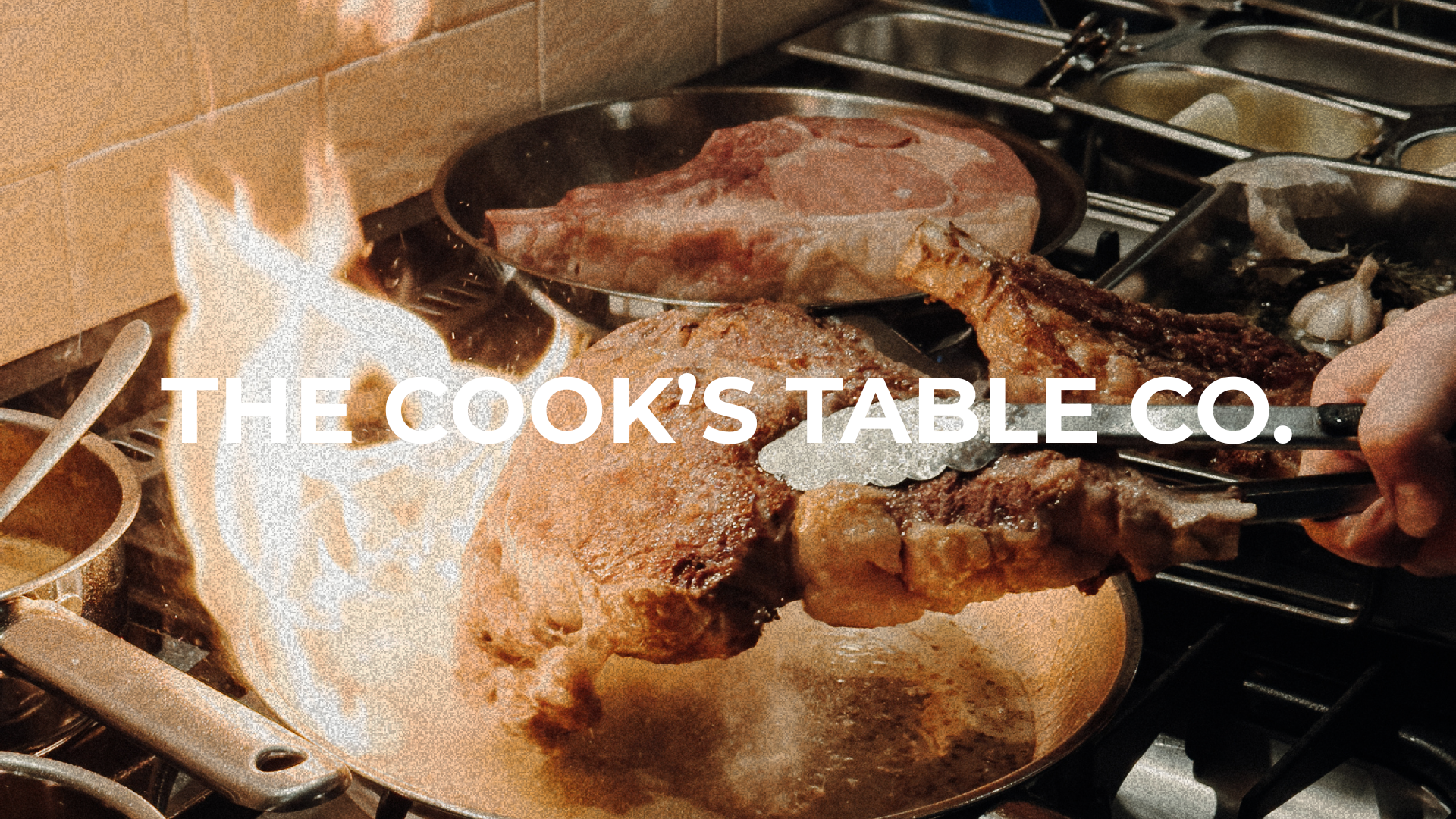Cooking steaks on a stove with flames visible, a hand using tongs to hold a large piece of meat, text overlay says 'The Cook's Table Co.'