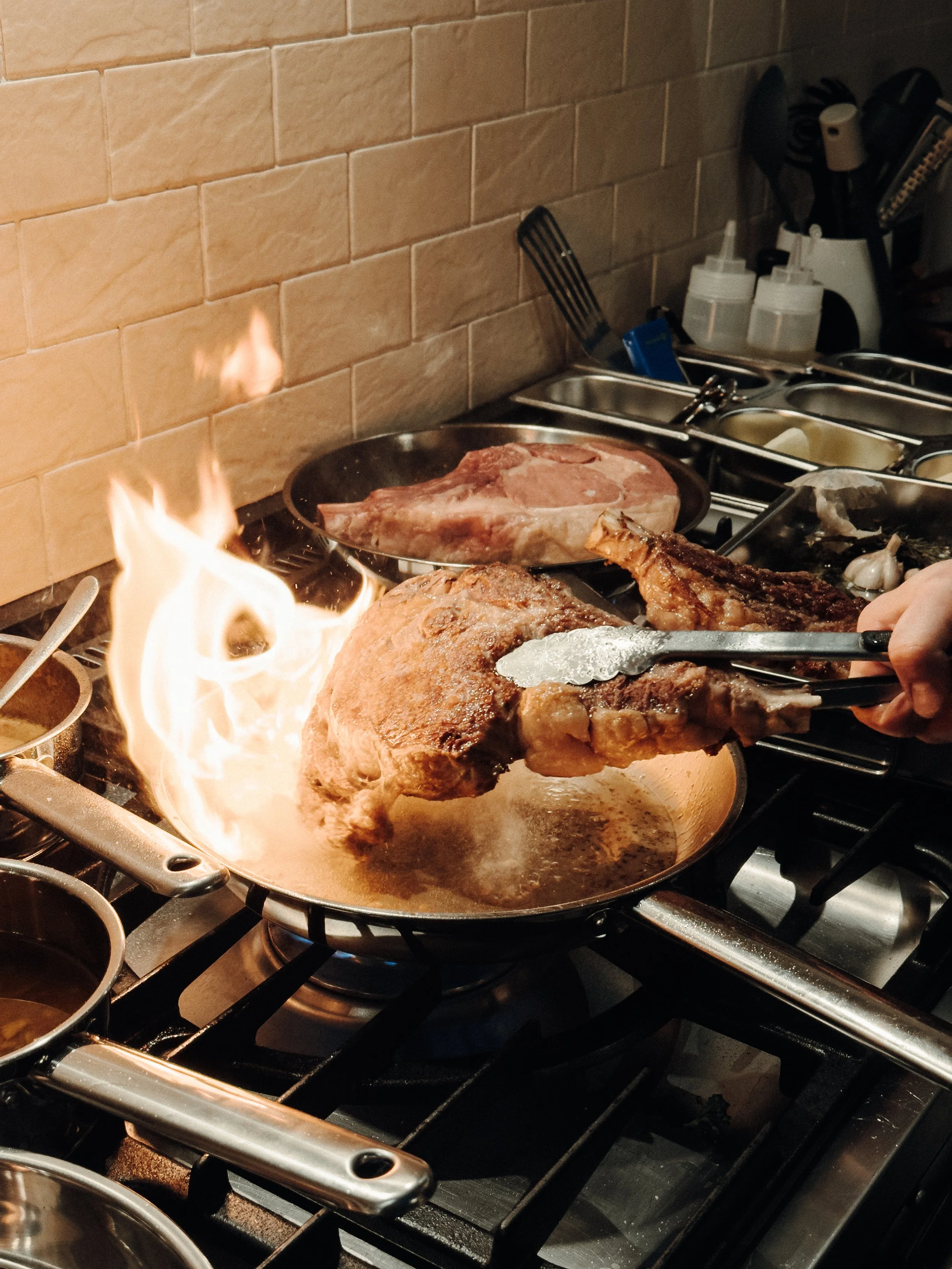 A large piece of beef being seared in a hot skillet with flames rising from it, in a professional kitchen. Various kitchen utensils and seasoning bottles are visible in the background.